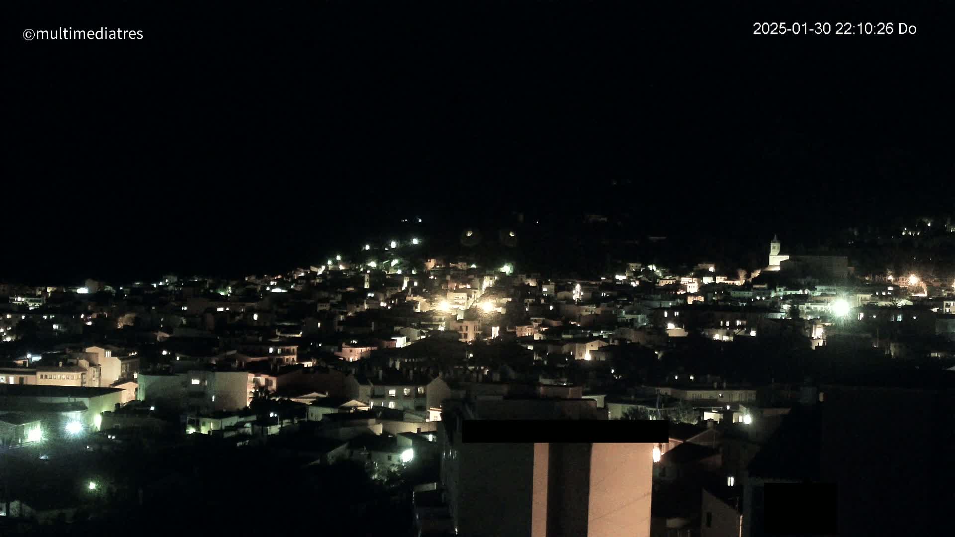 A nighttime, high-angle view of a town nestled on a hillside, with many buildings illuminated by lights.