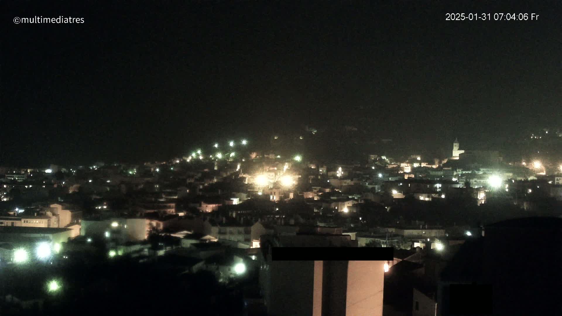 A nighttime view of a town nestled on a hillside, illuminated by numerous lights under a dark, possibly hazy sky.