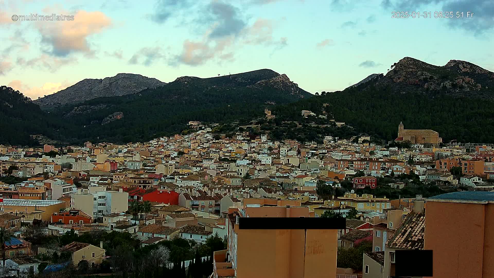 A sprawling town of low-rise buildings nestles at the foot of a mountain range under a partly cloudy sky.