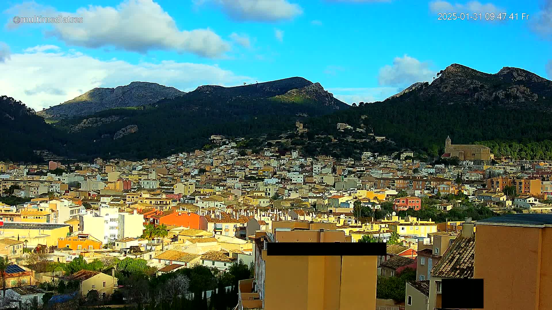 A sprawling town of low-rise buildings nestles at the foot of sunlit, verdant mountains under a partly cloudy blue sky.