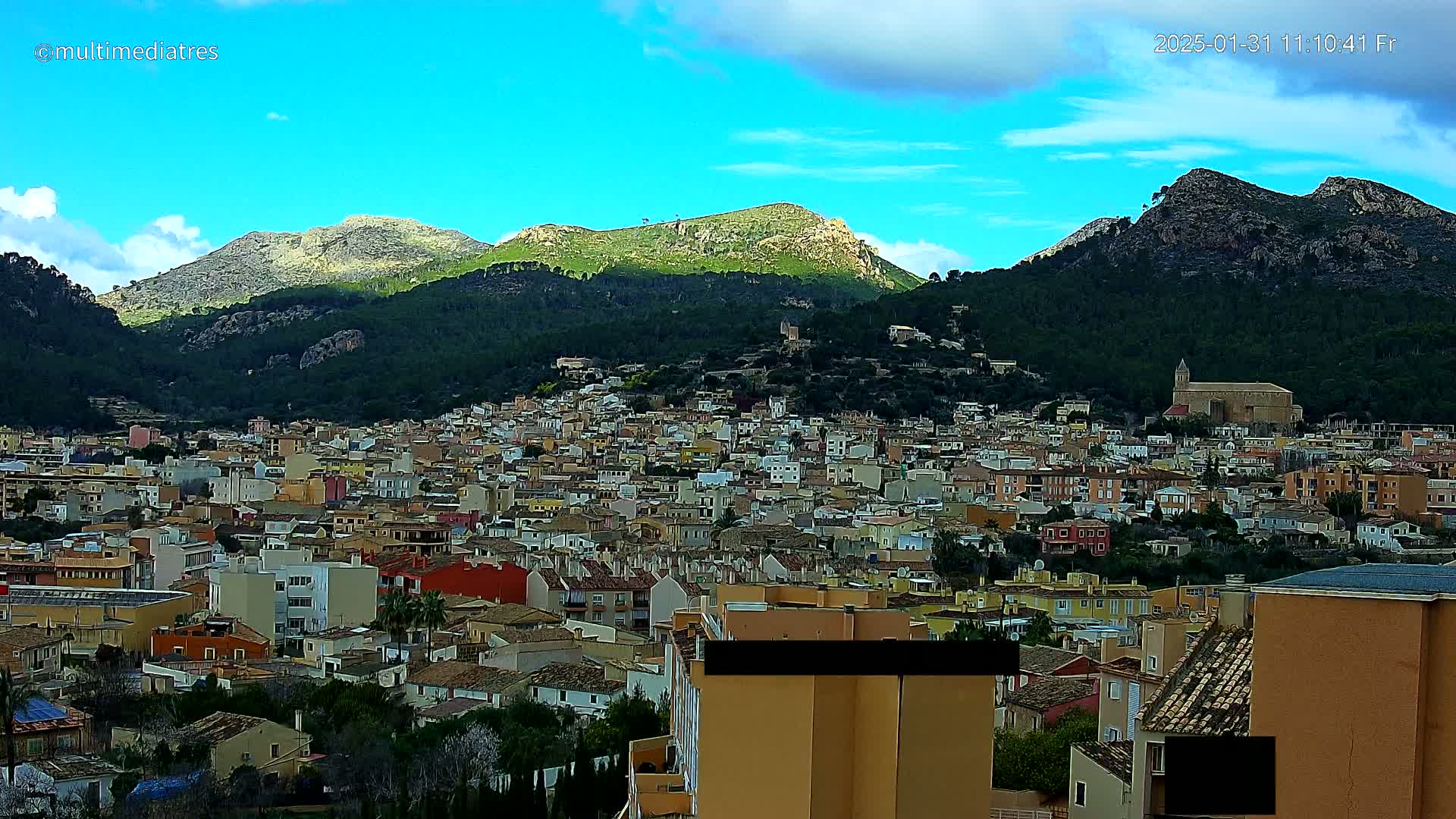 A sprawling town of closely packed buildings nestles at the base of verdant, rocky mountains under a partly cloudy, bright blue sky.