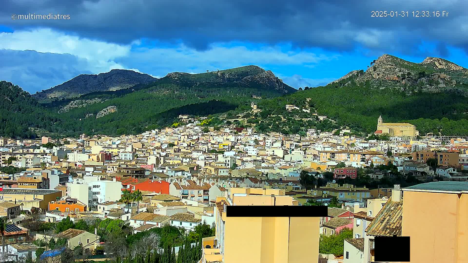 A sprawling town nestled at the base of verdant, rocky mountains under a partly cloudy sky.