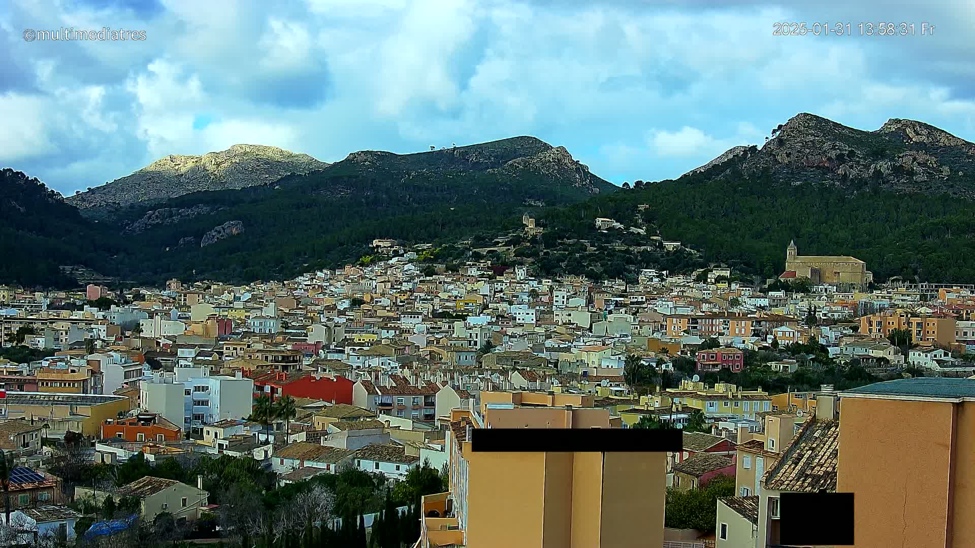 A sprawling town of colorful buildings nestles at the base of a large, verdant mountain range under a partly cloudy sky.