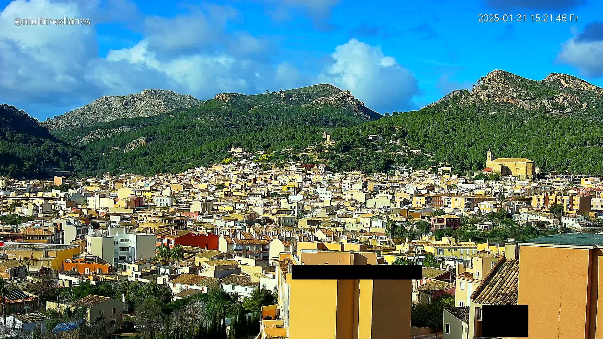 A sprawling town of light-colored buildings nestled in a valley, surrounded by lush green hills under a mostly sunny sky with some clouds.