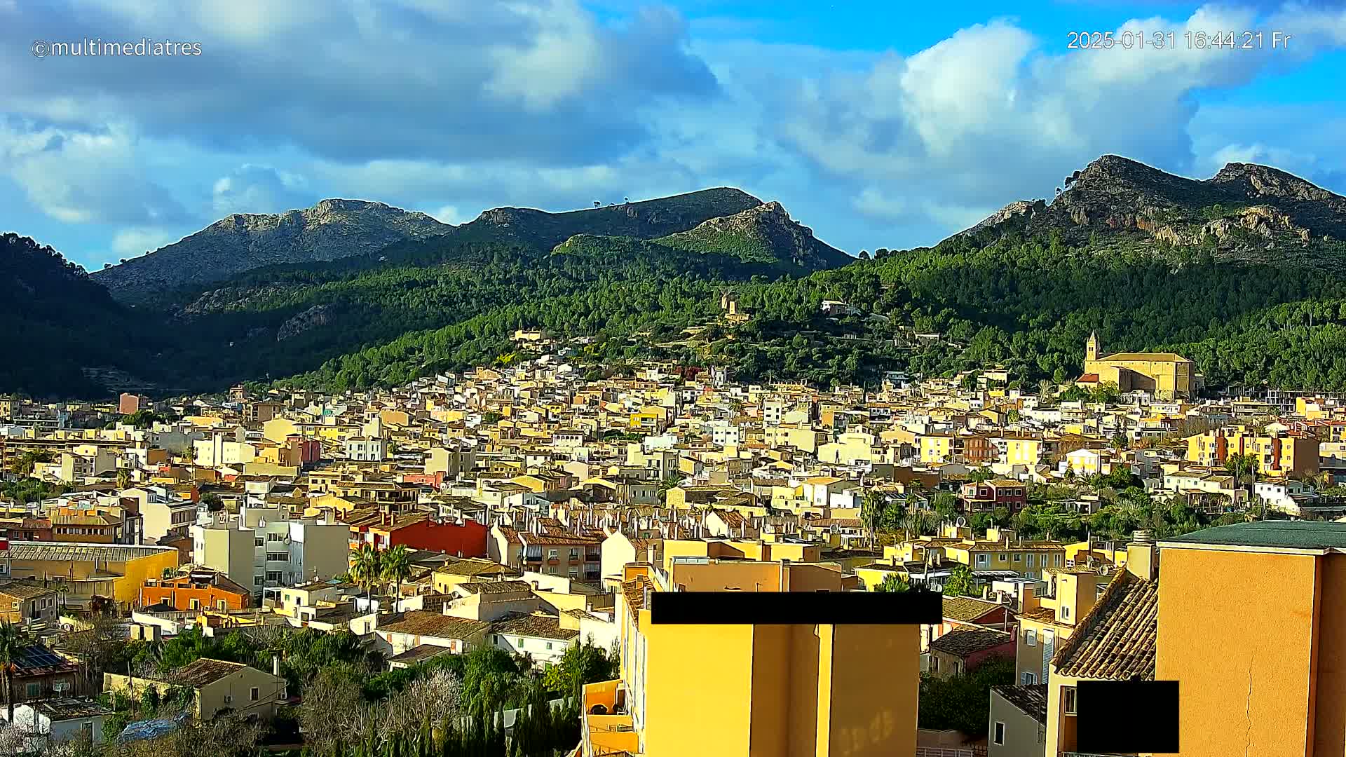 A sprawling town of light-colored buildings nestled at the base of a verdant mountain range under a partly cloudy sky.