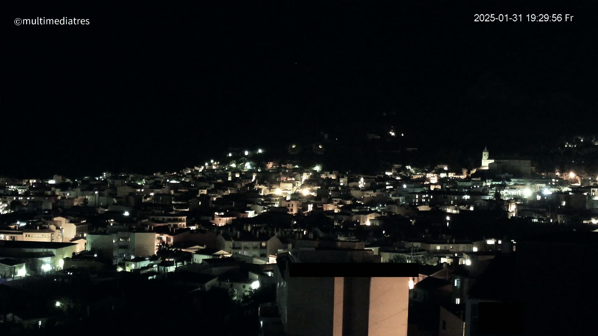 A nighttime view of a town nestled on a hillside, illuminated by scattered lights under a clear, dark sky.