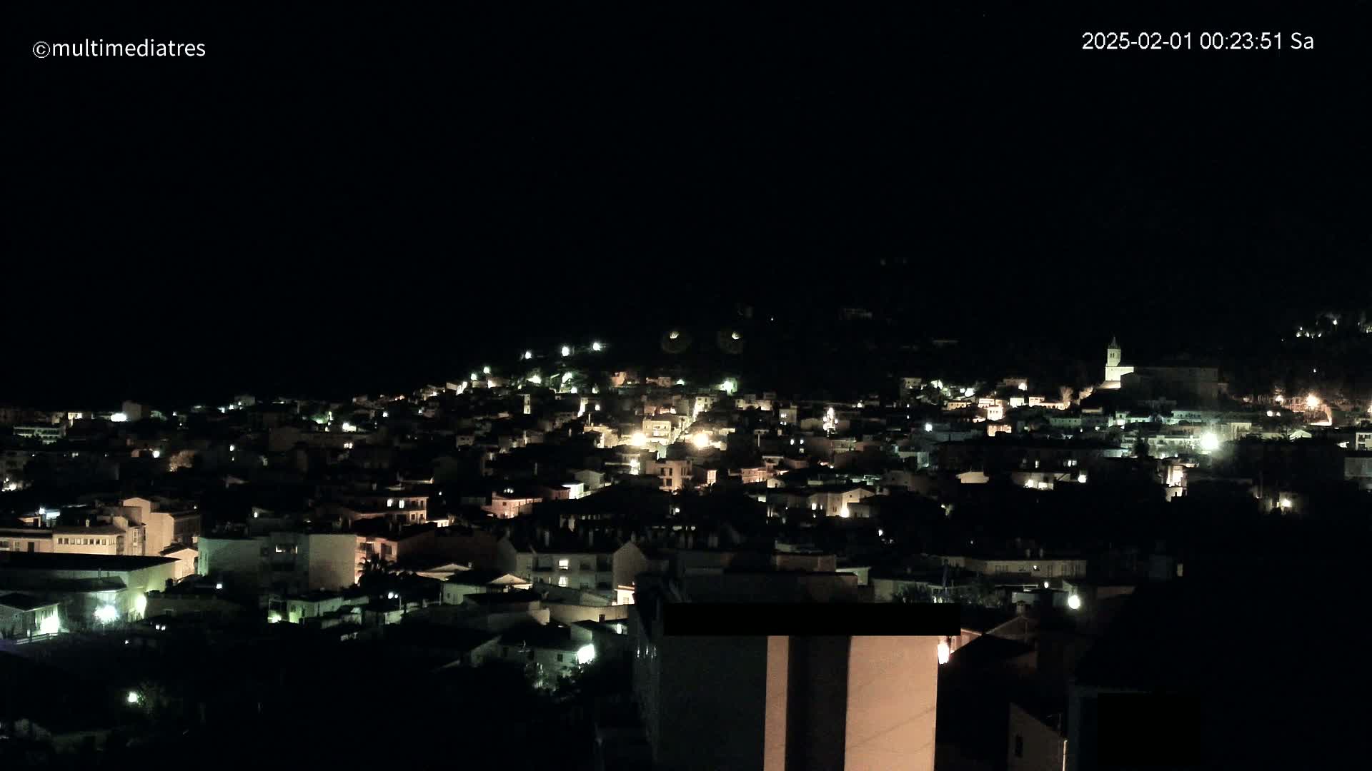 A nighttime view of a town nestled on a hillside, with scattered lights illuminating buildings under a clear, dark sky.