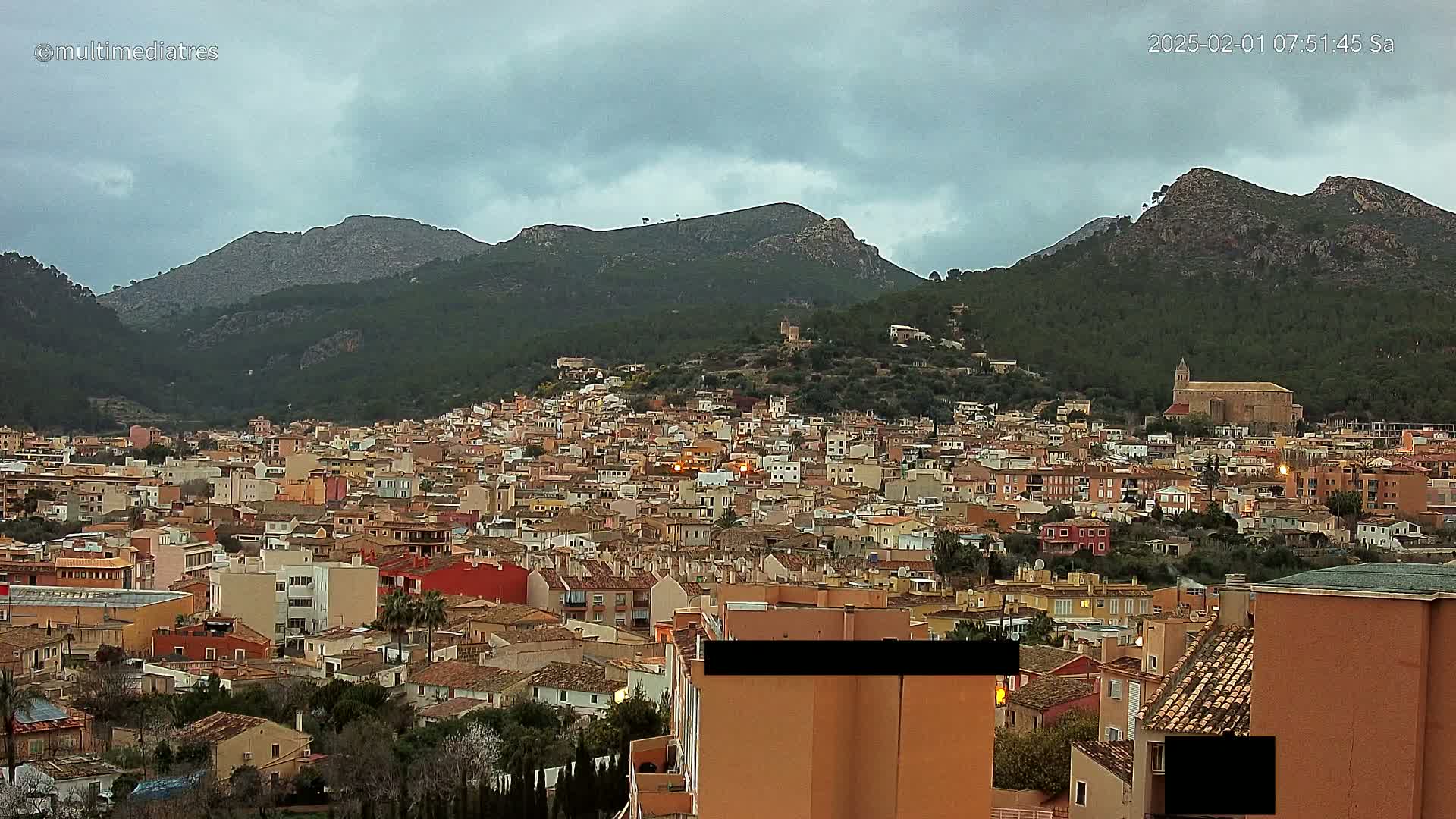 A sprawling town of tan and orange buildings nestled in a valley between dark green, mountainous hills under a cloudy sky.