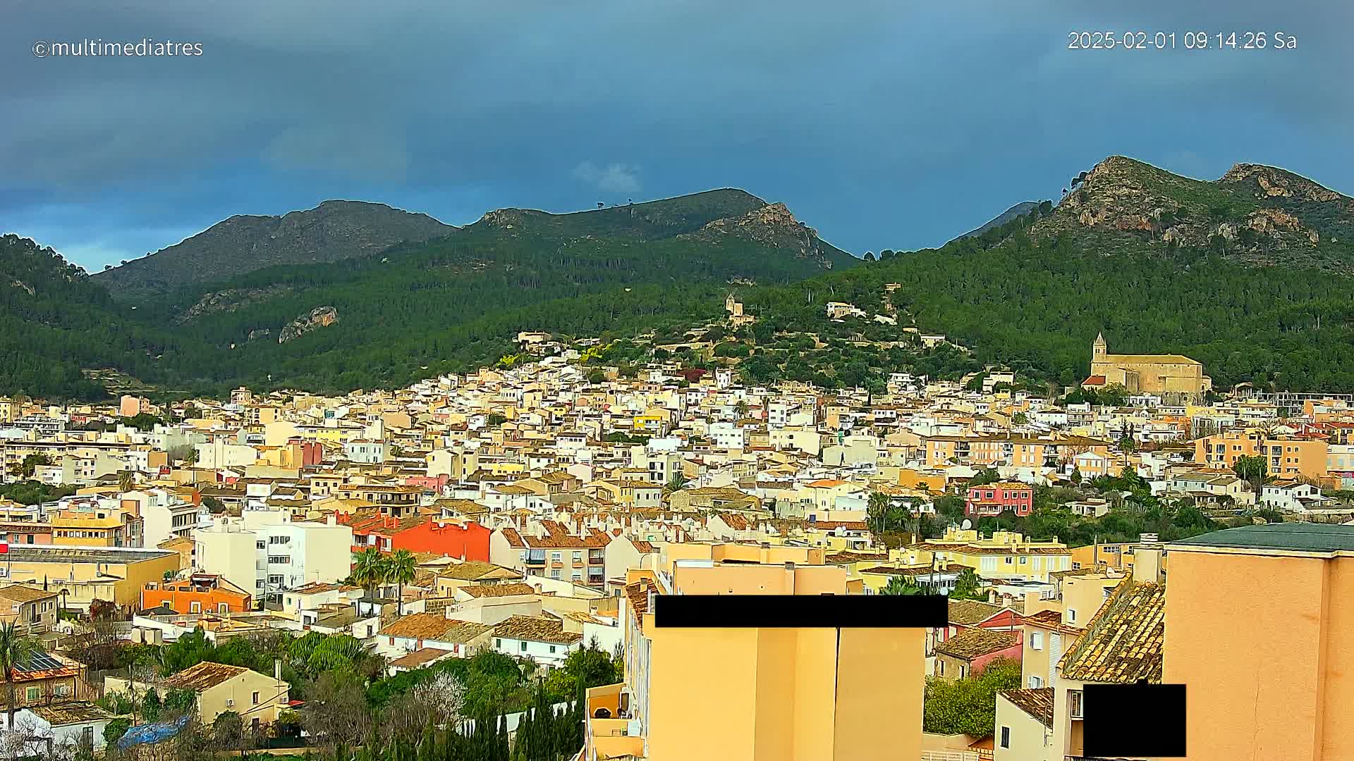 A sprawling town of light-colored buildings nestles in a valley surrounded by green, tree-covered mountains under a mostly cloudy sky.