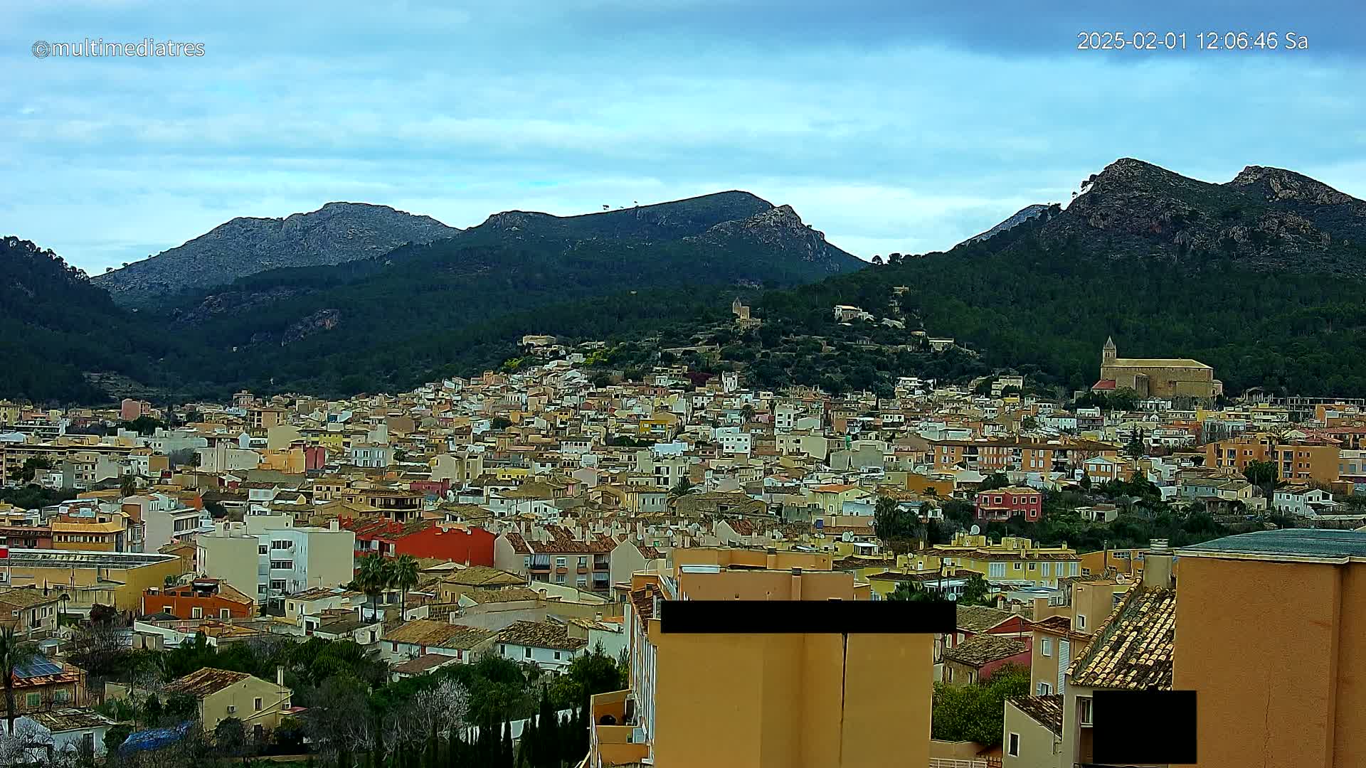 A sprawling town of closely packed, colorful buildings nestles at the base of verdant, rocky mountains under a mostly clear, light blue sky.