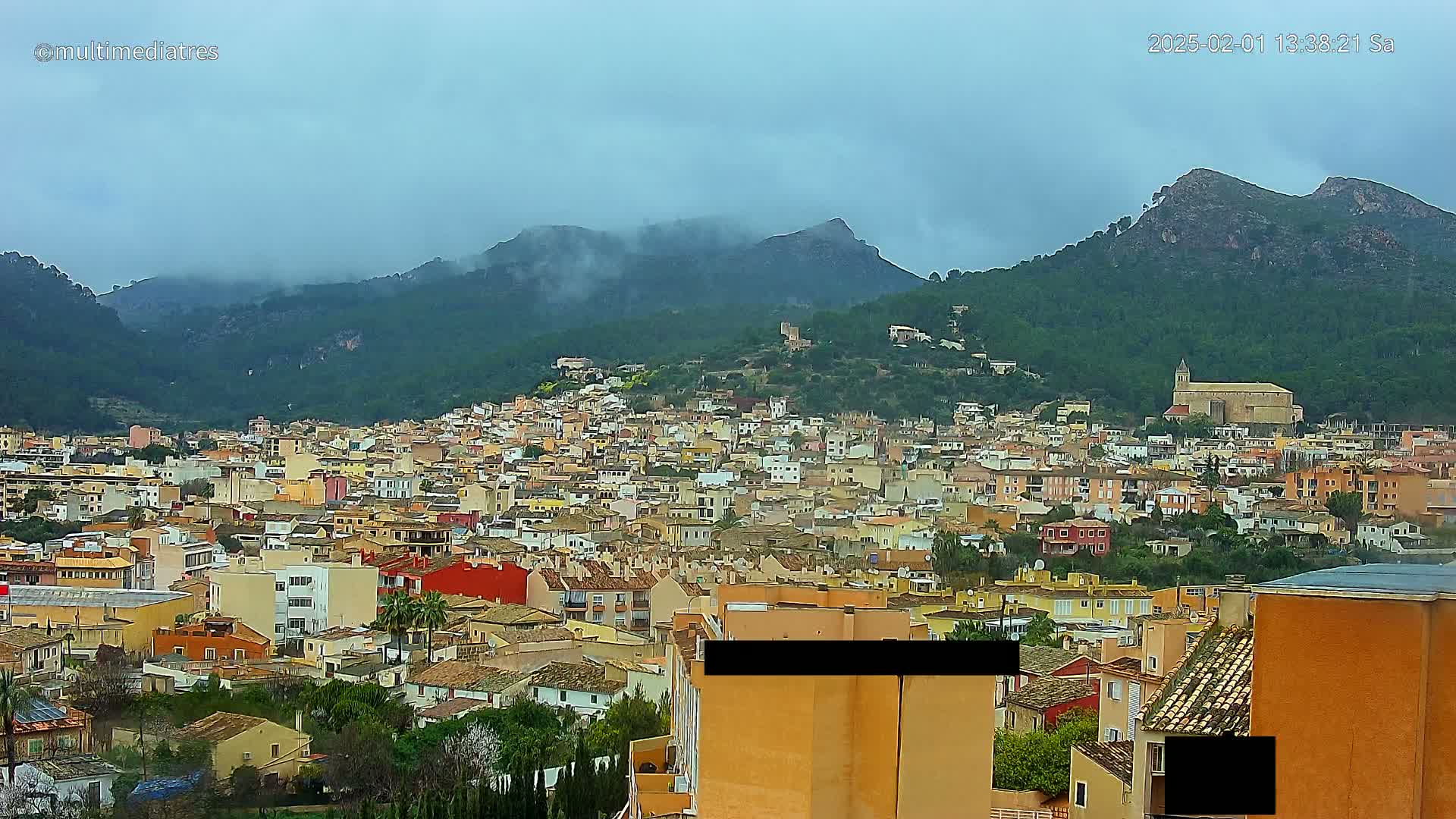 A dense collection of buildings nestled in a valley, overlooked by mountains shrouded in mist under a cloudy sky.