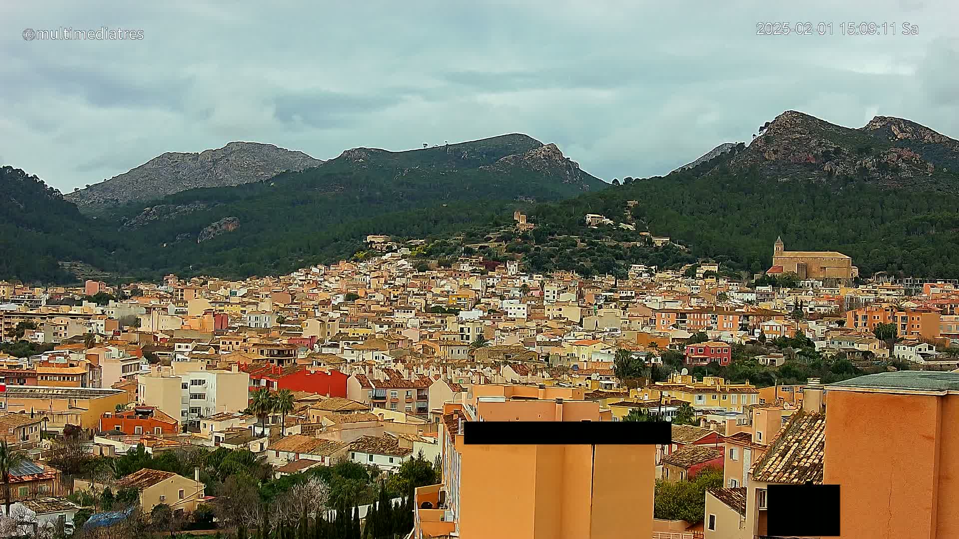 A sprawling town of low-rise buildings nestled in a valley, backed by verdant mountains under a cloudy sky.