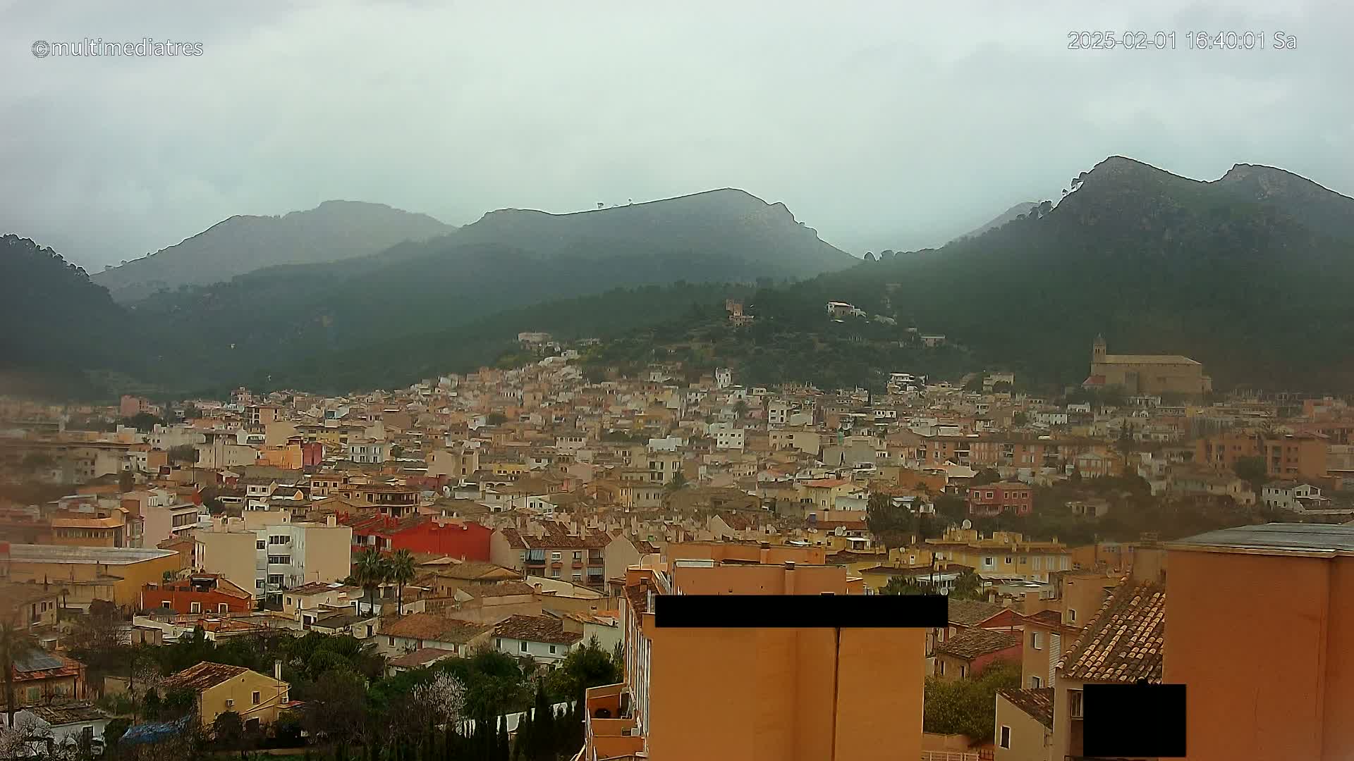A dense collection of low-rise buildings nestled in a valley, surrounded by green, mountainous terrain under an overcast sky.