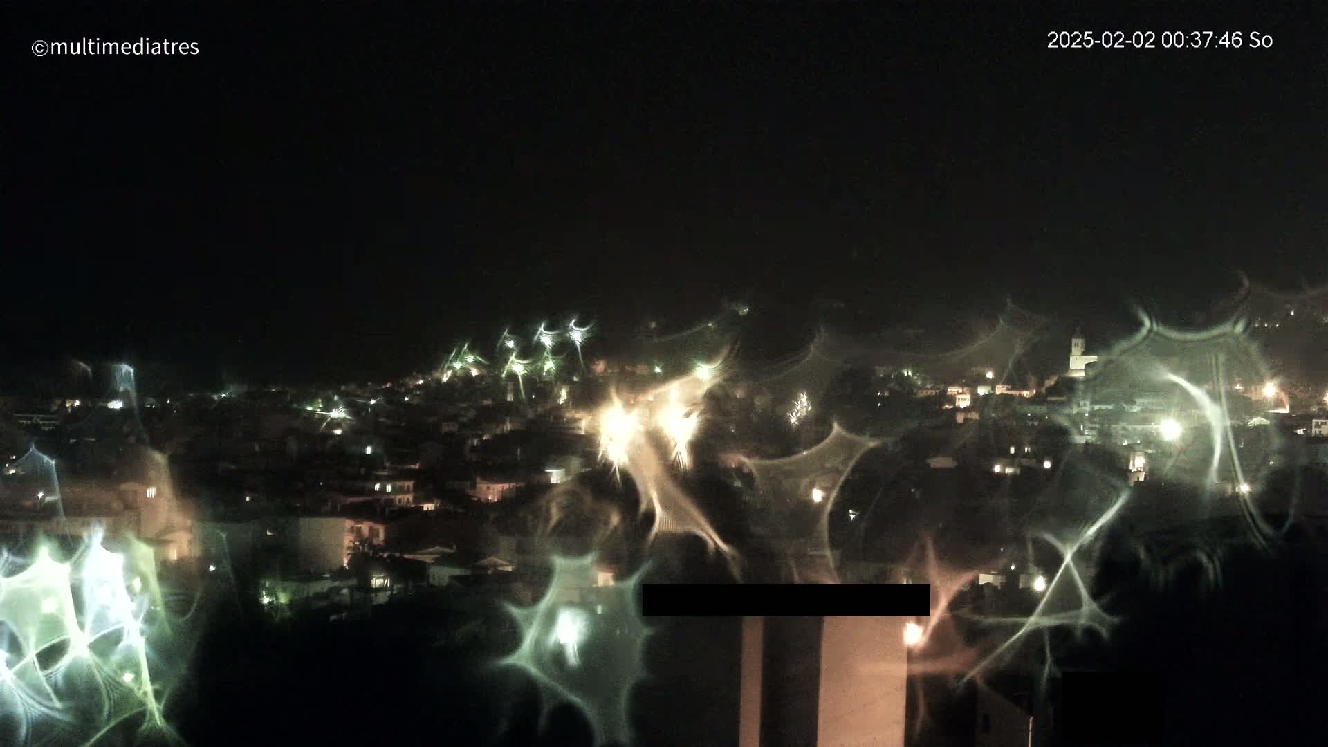 A nighttime view of a town illuminated by lights, seen through a window with distorted reflections creating a hazy, streaked effect.