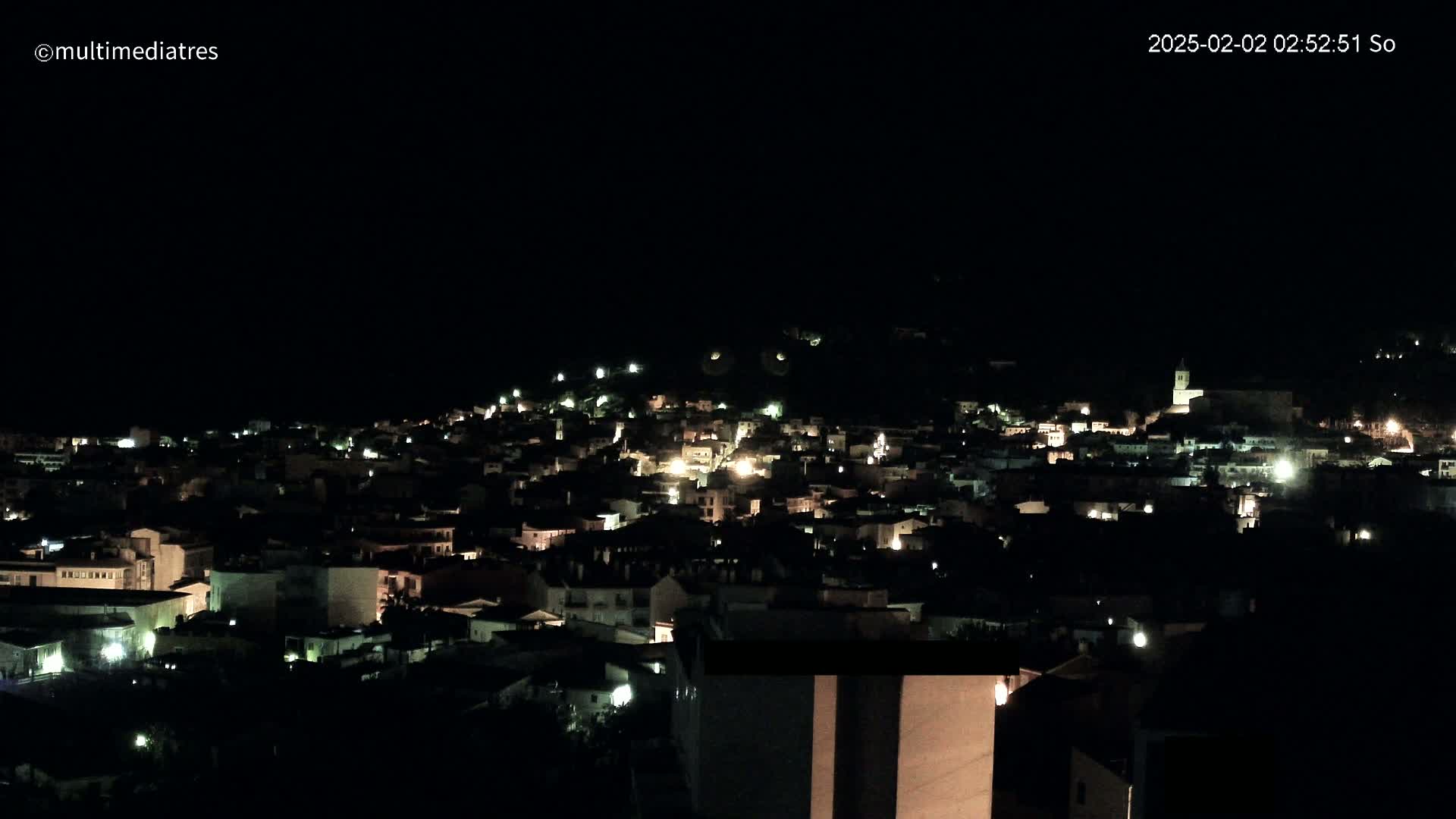 A nighttime, high-angle view of a town nestled on a hill, with many windows illuminated.