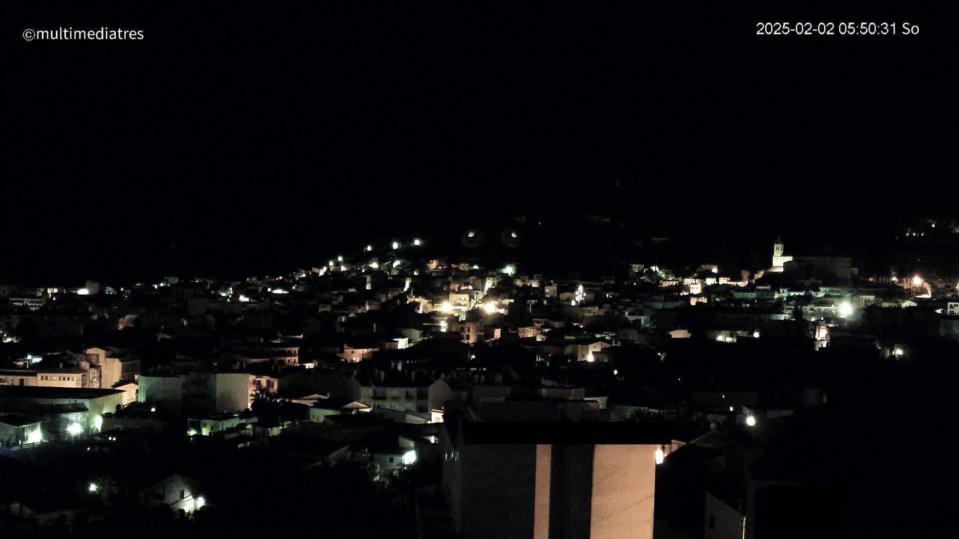 A nighttime view of a town nestled on a hill, with scattered lights illuminating the buildings under a dark, clear sky.