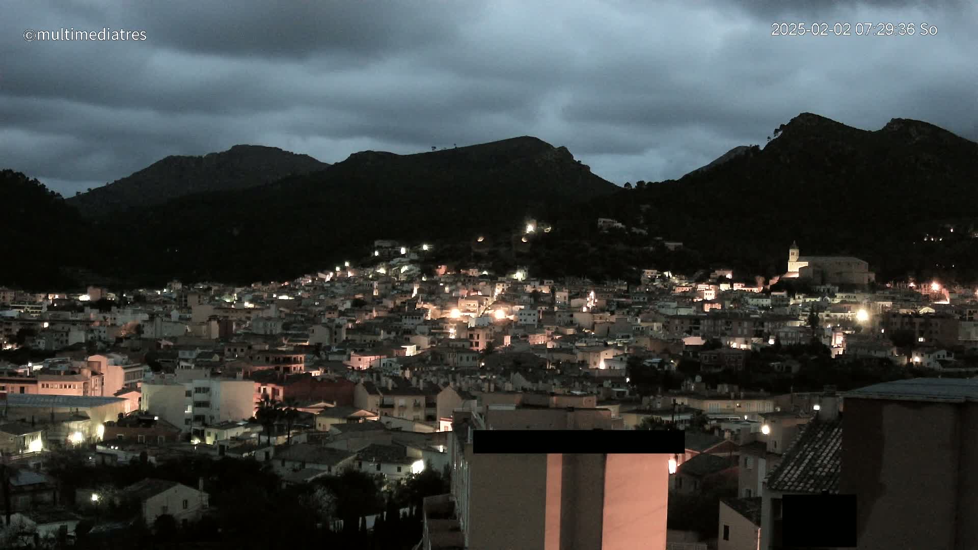 A nighttime view of a town nestled in a valley between dark mountains under a cloudy sky.