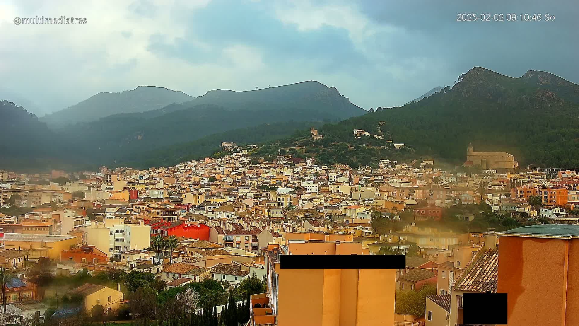 A sprawling town of tan and orange buildings nestles at the base of a mountain range under a cloudy sky.
