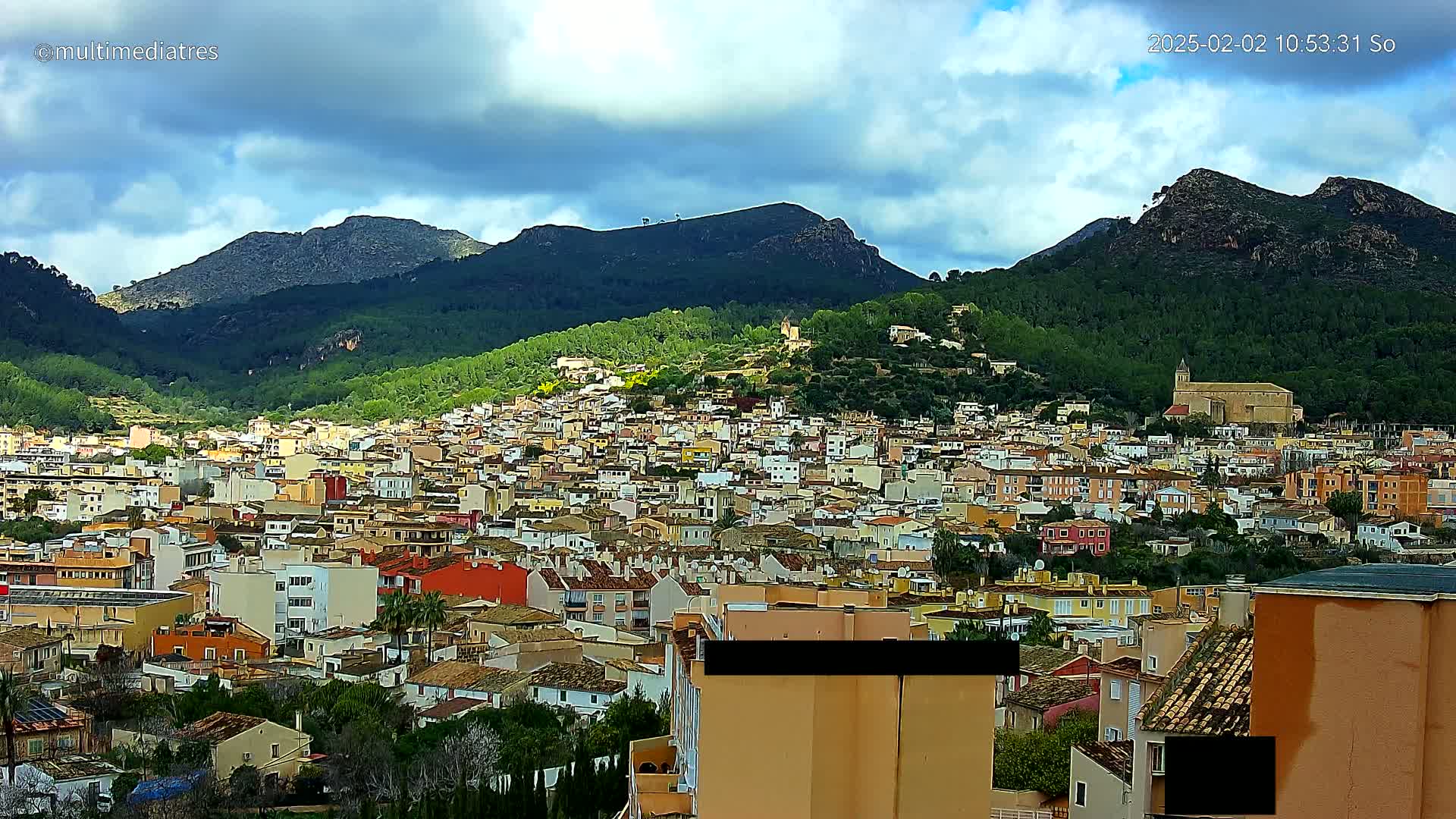 A sprawling town of light-colored buildings nestled in a valley between verdant, mountainous terrain under a partly cloudy sky.