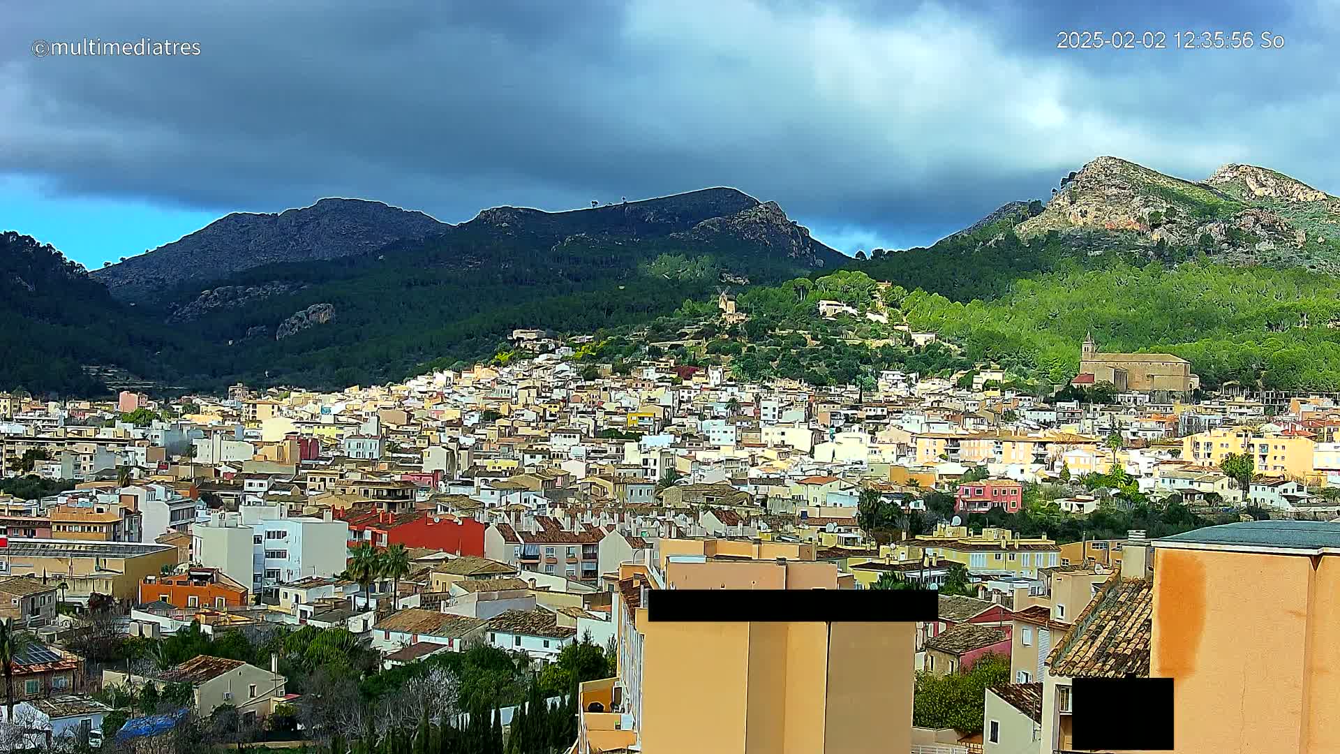 A sprawling town of low-rise buildings nestles at the base of a mountainous region under a partly cloudy sky.