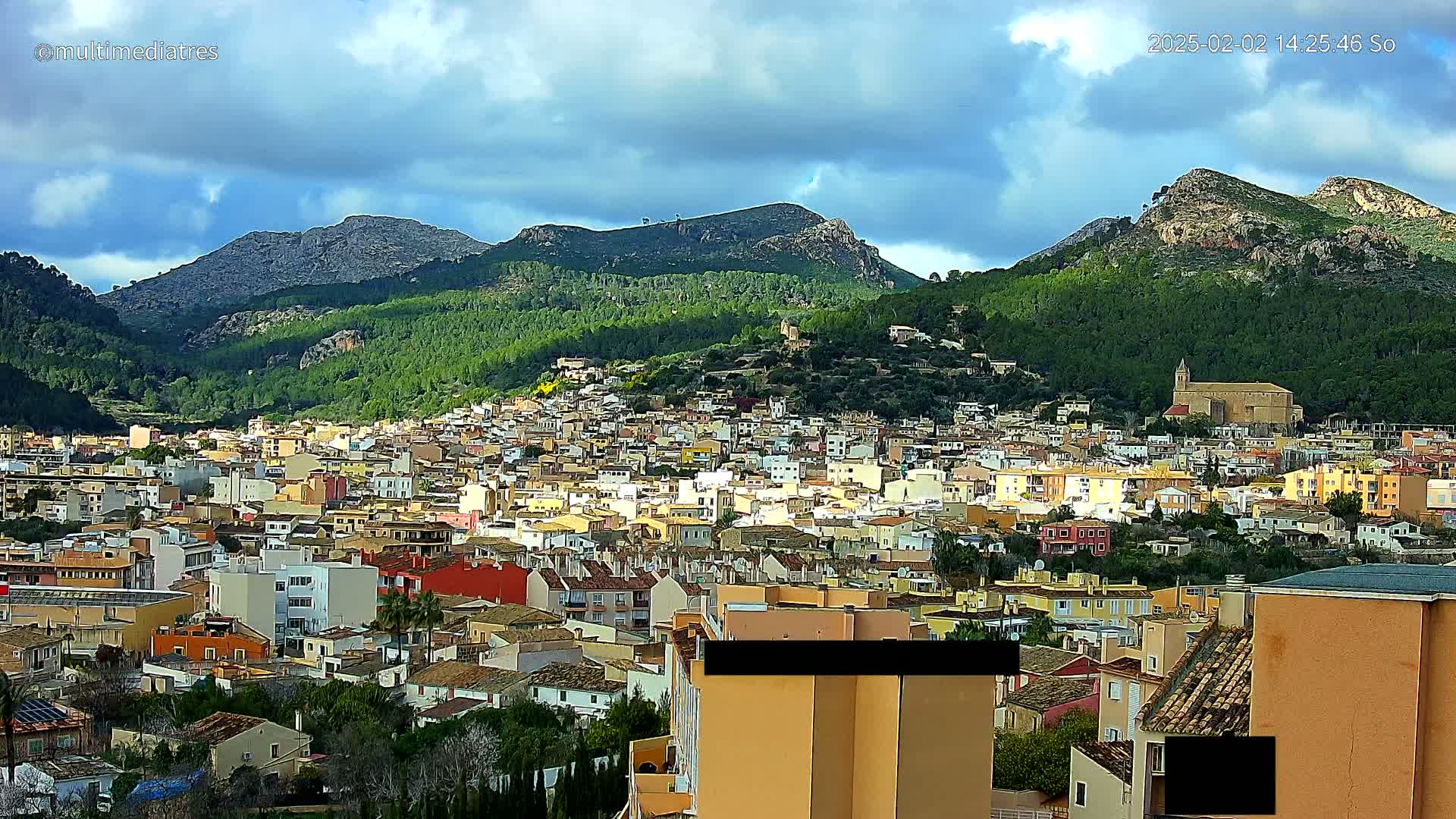 A sprawling town of closely packed buildings nestles in a valley surrounded by steep, green-covered mountains under a partly cloudy sky.