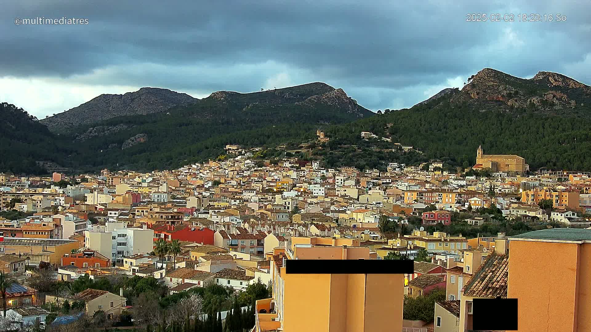A sprawling town of low-rise buildings nestles at the foot of forested, mountainous terrain under a cloudy sky.