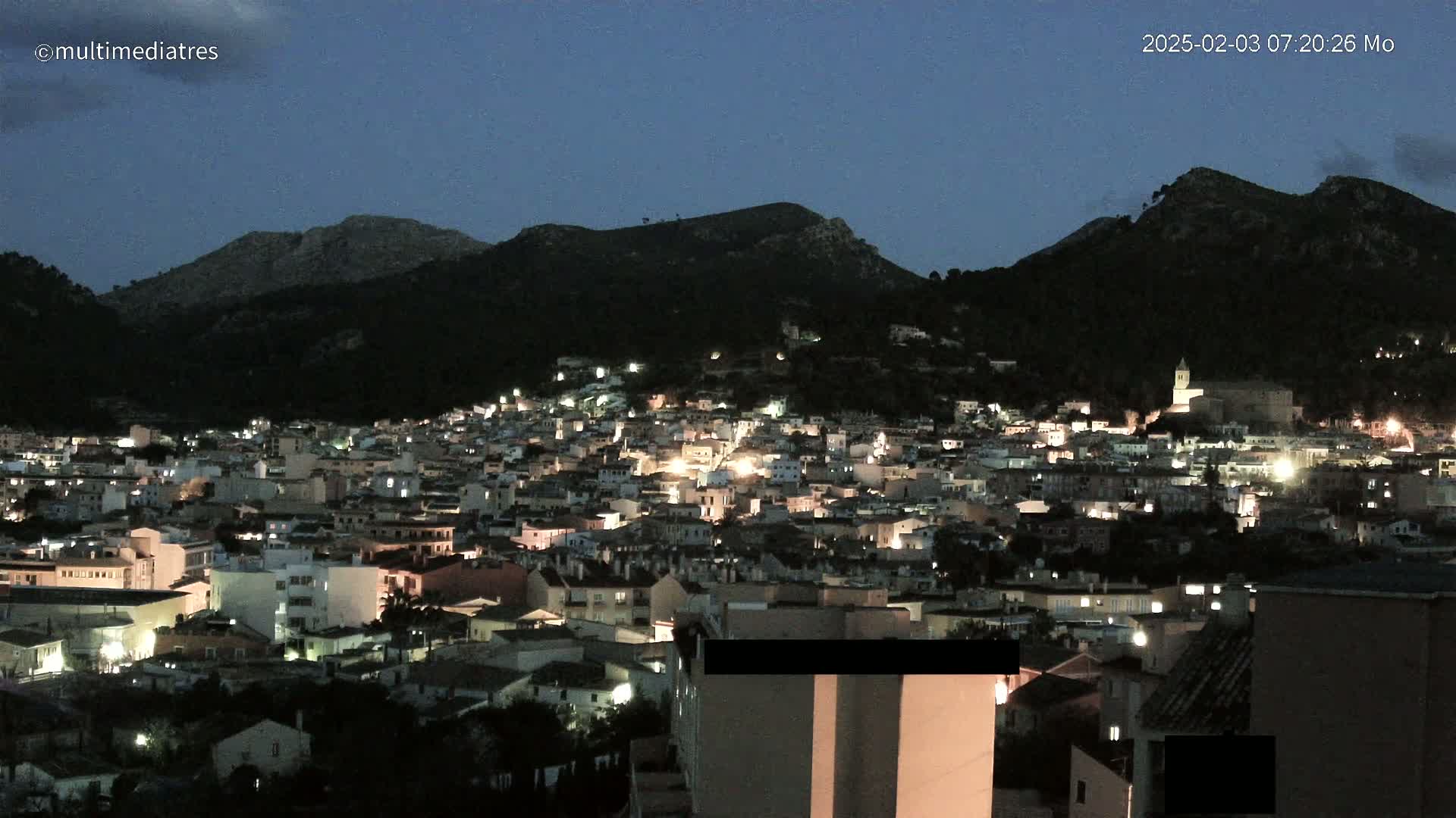 A nighttime view of a town nestled in a valley between dark hills under a twilight sky.