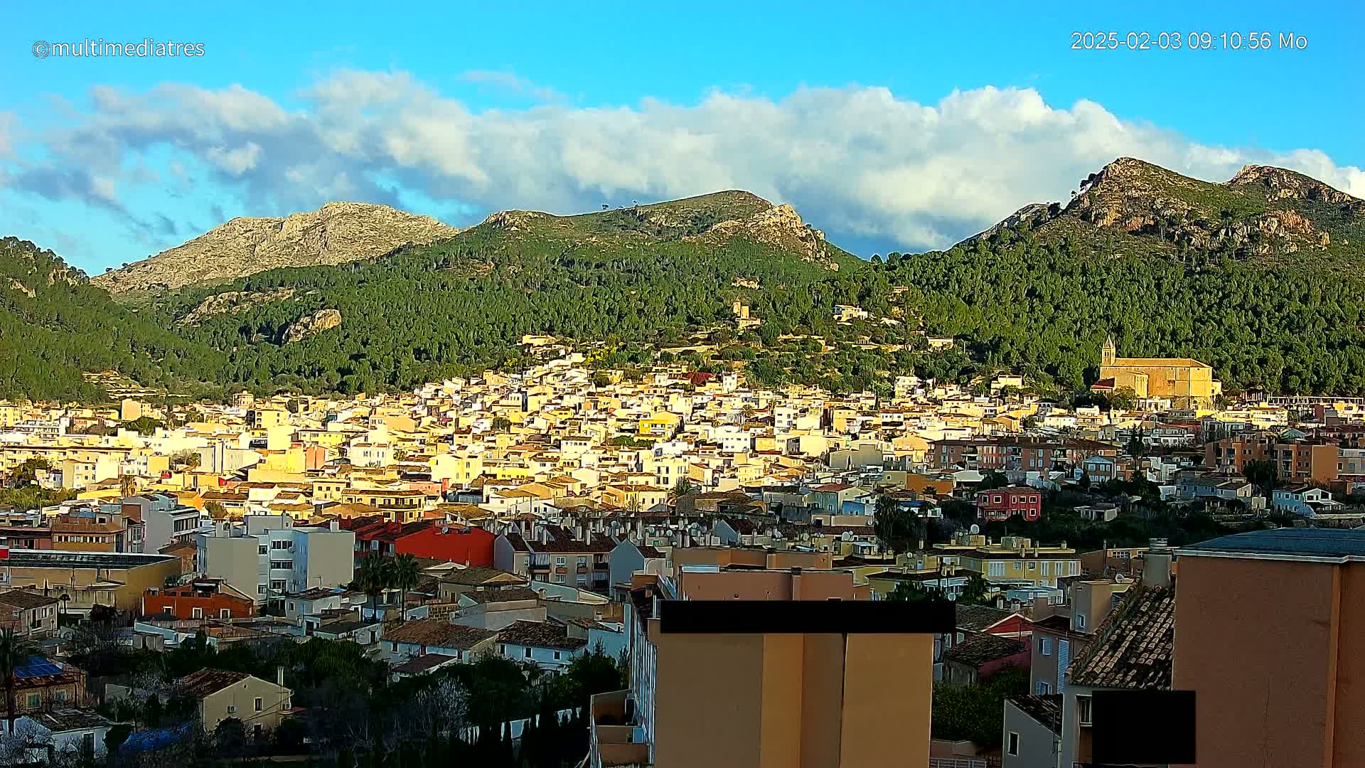 A sprawling town of light-yellow buildings nestled in a lush green valley, partially surrounded by rocky, tree-covered mountains under a mostly sunny sky with some scattered clouds.