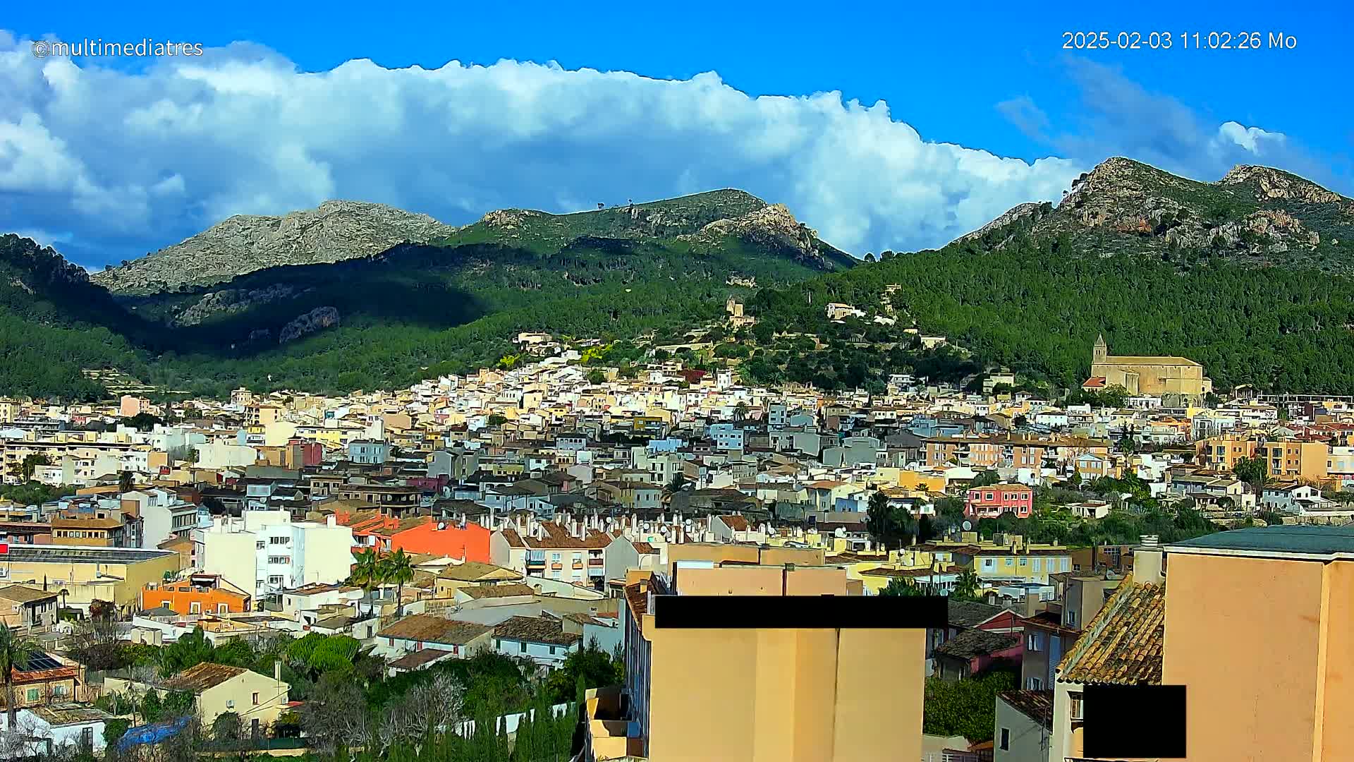 A sprawling town of low-rise buildings nestles at the base of a verdant mountain range under a partly cloudy blue sky.