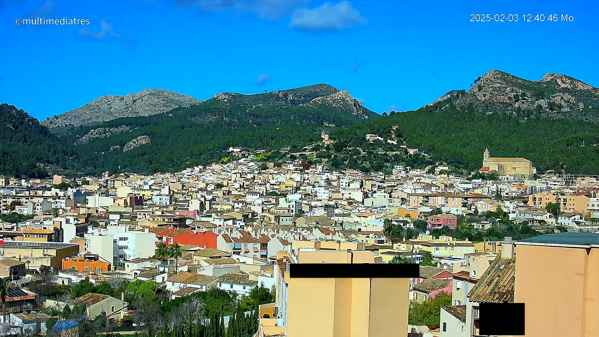 A sprawling town of light-colored buildings nestles at the base of lush green mountains under a clear blue sky.
