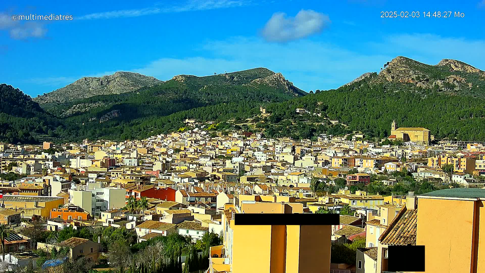 A sprawling town of low-rise buildings nestles at the base of verdant, rocky mountains under a clear blue sky.