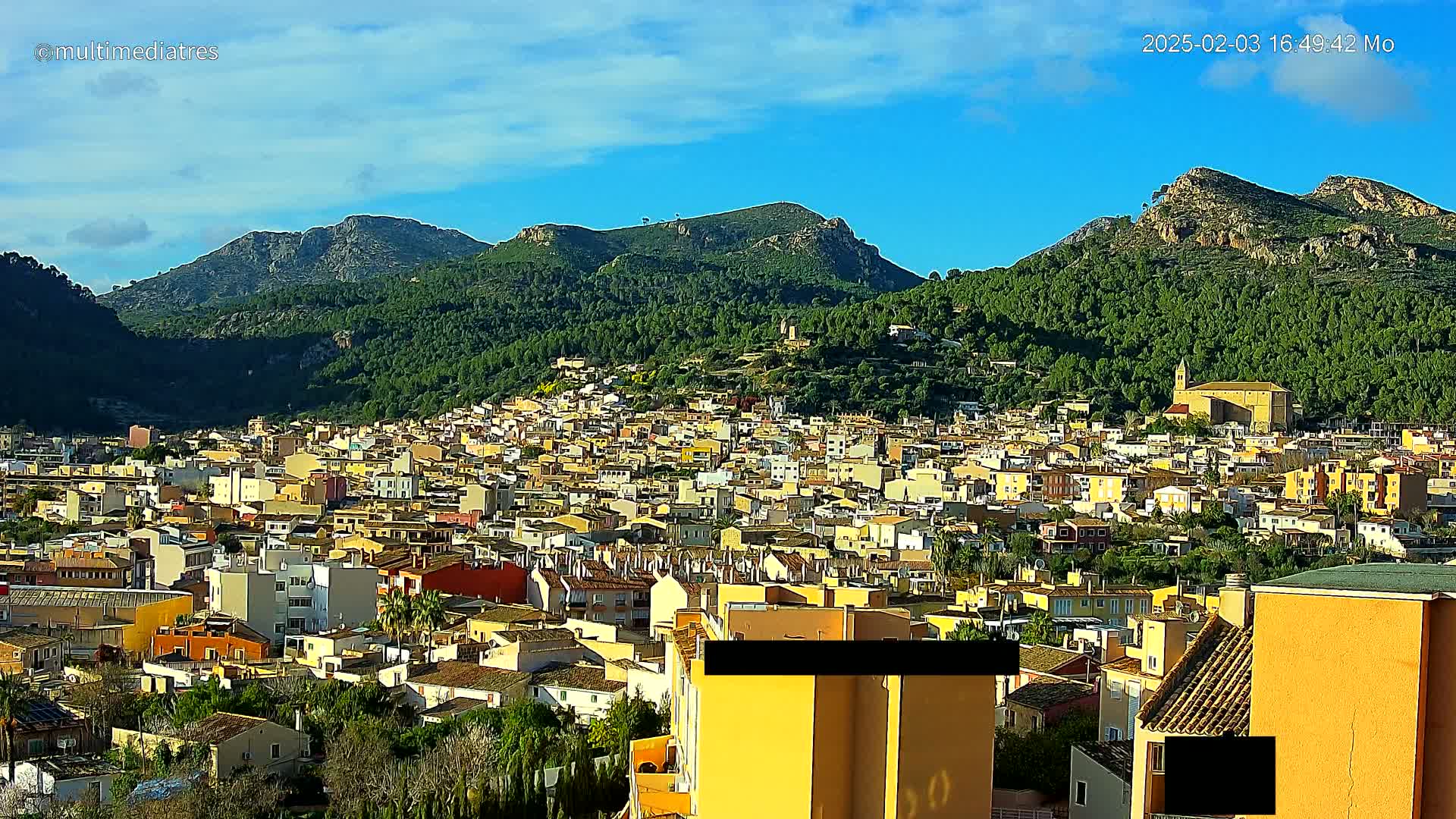 A sprawling town of low-rise buildings nestles in a valley surrounded by green, mountainous terrain under a mostly clear, sunny sky.