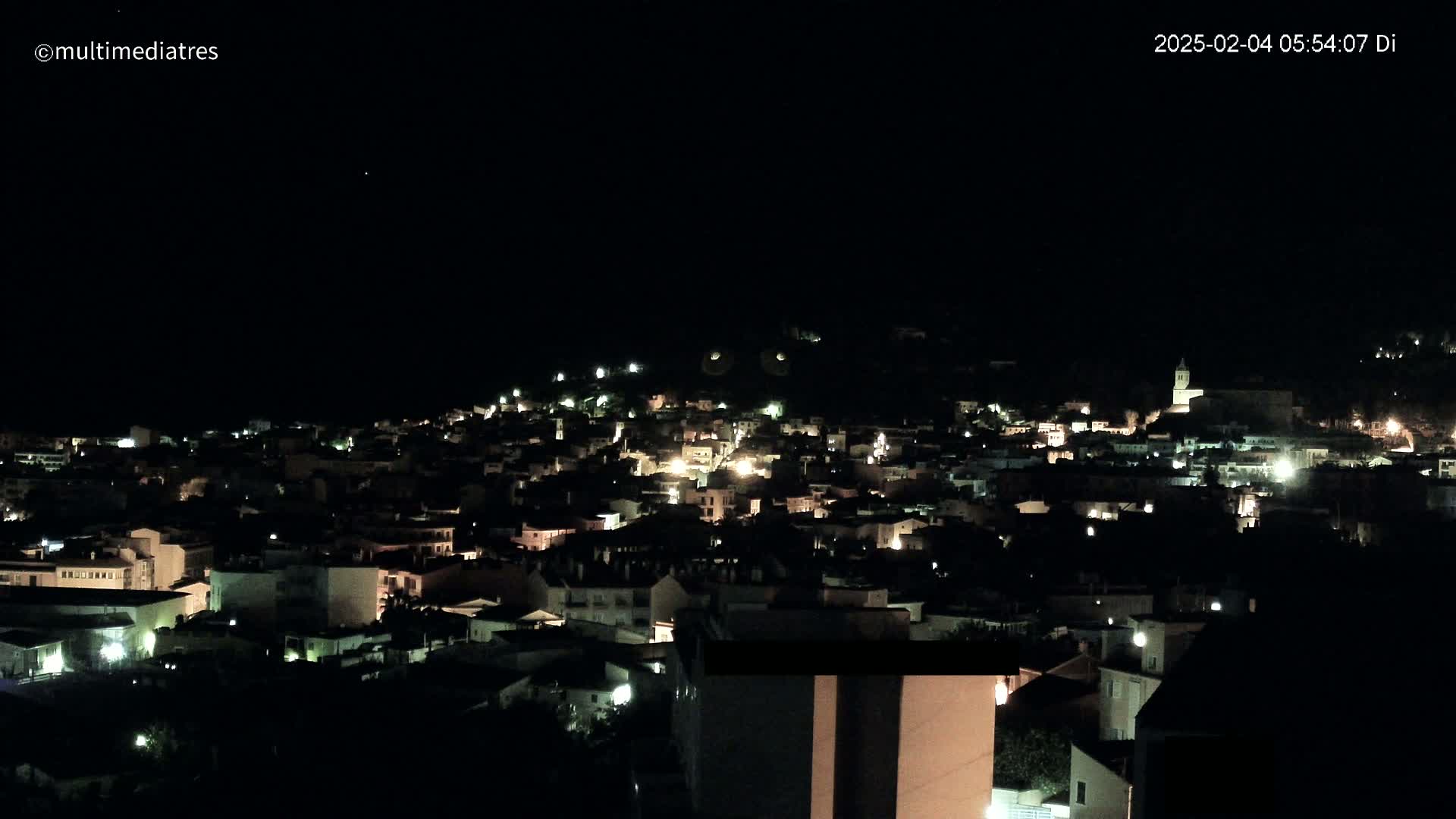 A nighttime, high-angle view shows a town nestled on a hillside, its buildings illuminated by scattered lights.