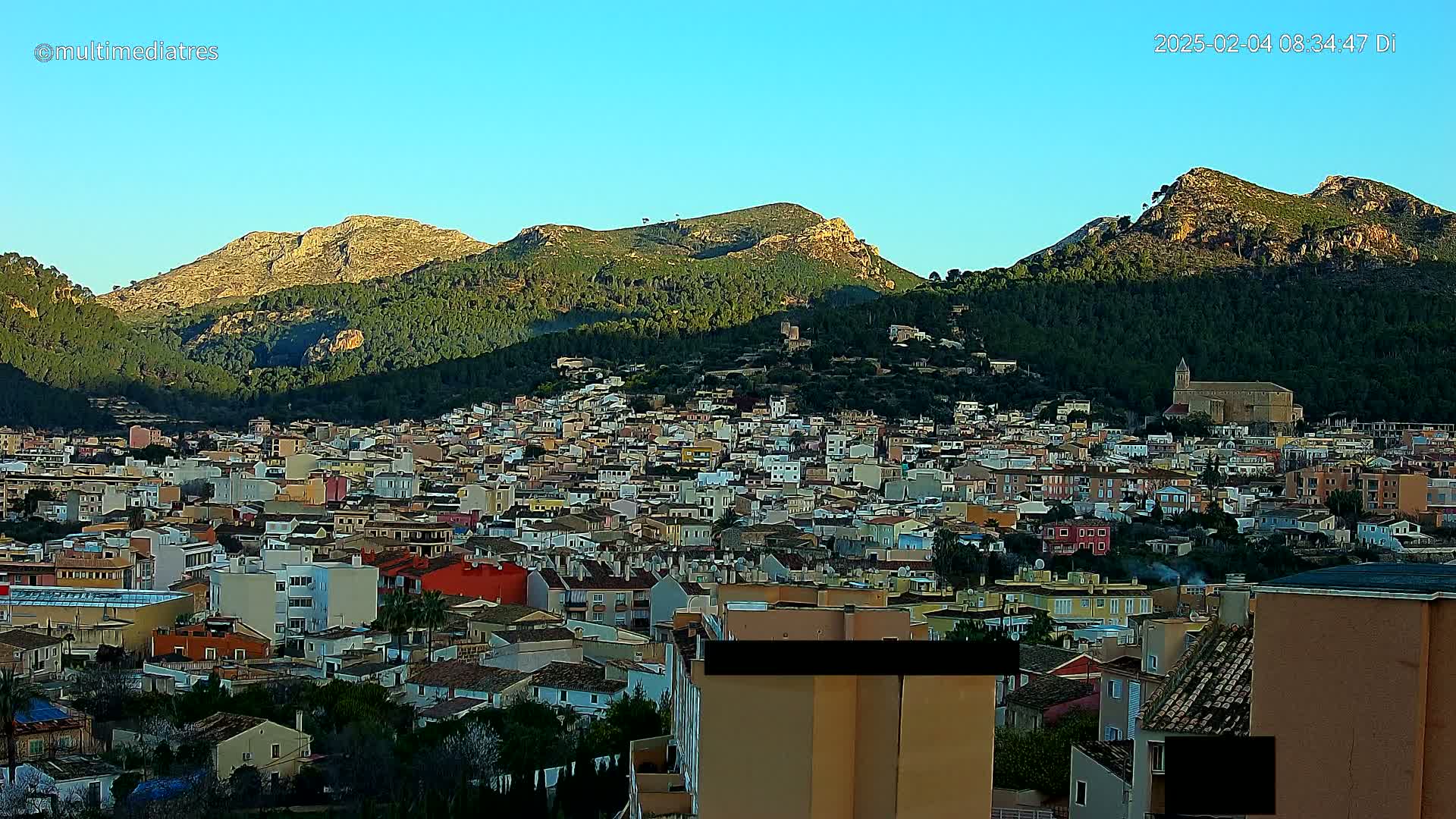 A sprawling town of closely packed buildings nestled in a valley, surrounded by verdant hills and mountains under a clear blue sky.