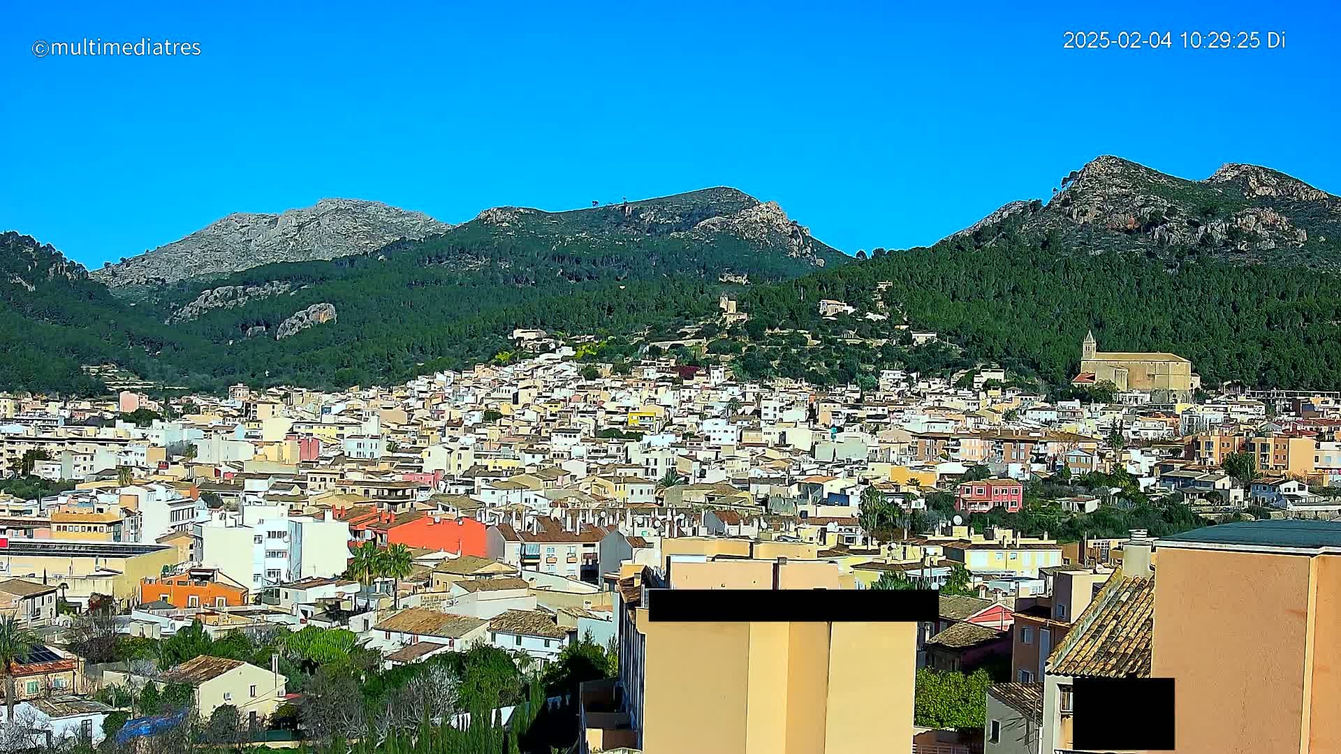 A sprawling town of light-colored buildings nestles at the foot of verdant, rocky mountains under a clear blue sky.