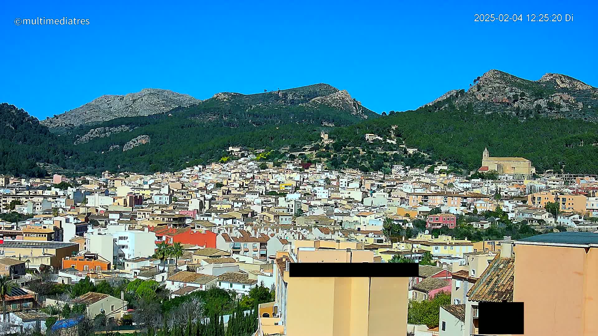 A sprawling town of low-rise buildings nestles at the foot of a verdant mountain range under a clear blue sky.