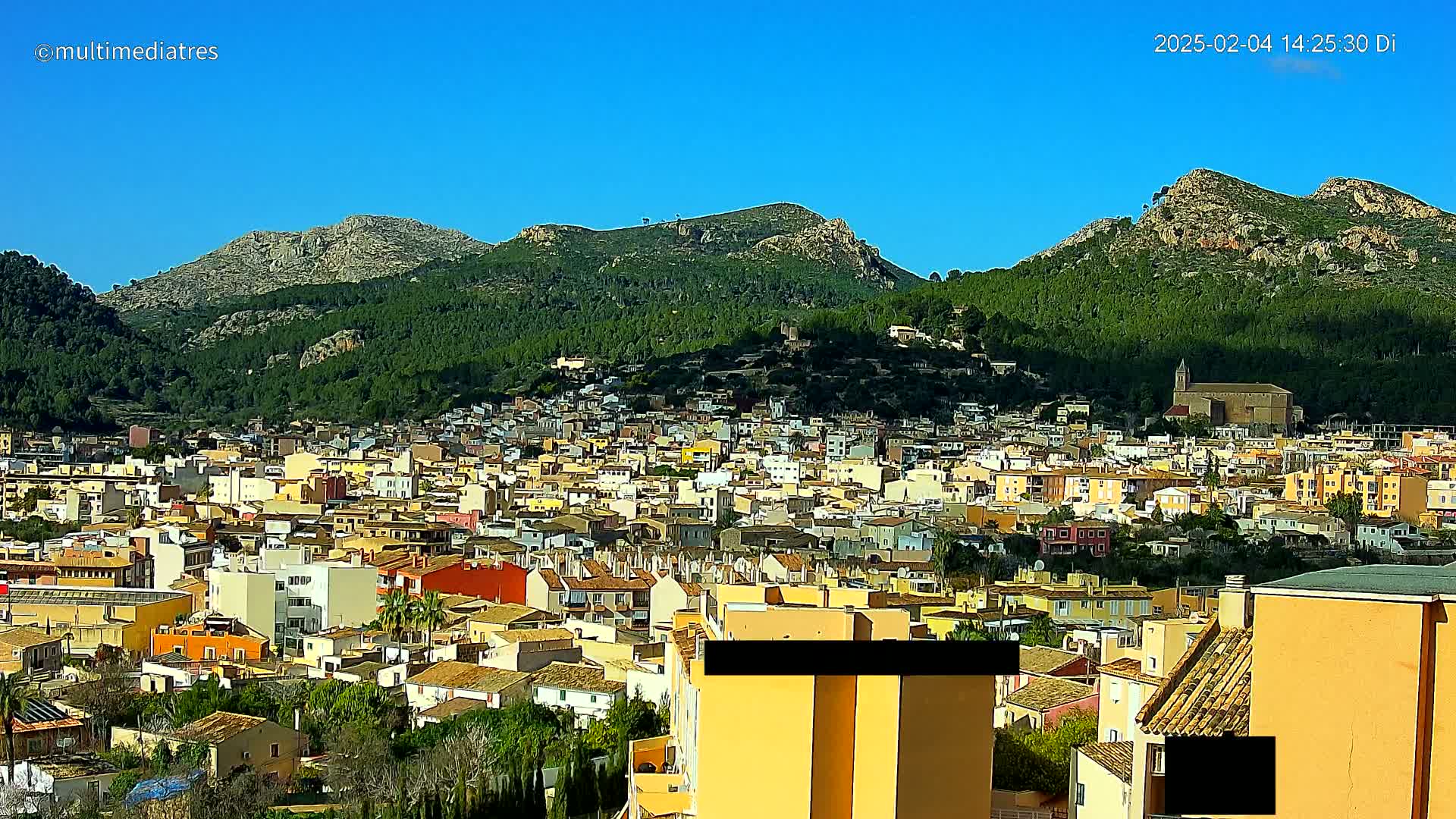A sprawling town of low-rise buildings nestles at the base of a verdant mountain range under a clear blue sky.