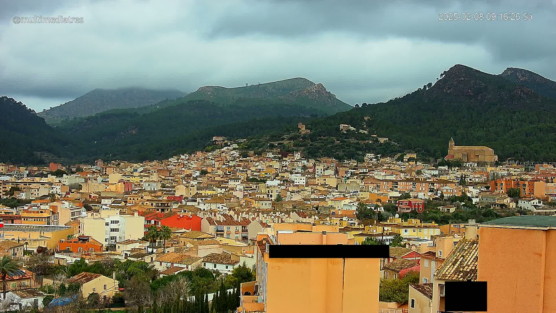 A sprawling town of predominantly low-rise, tan buildings nestled in a valley between verdant, mountainous terrain under a cloudy sky.