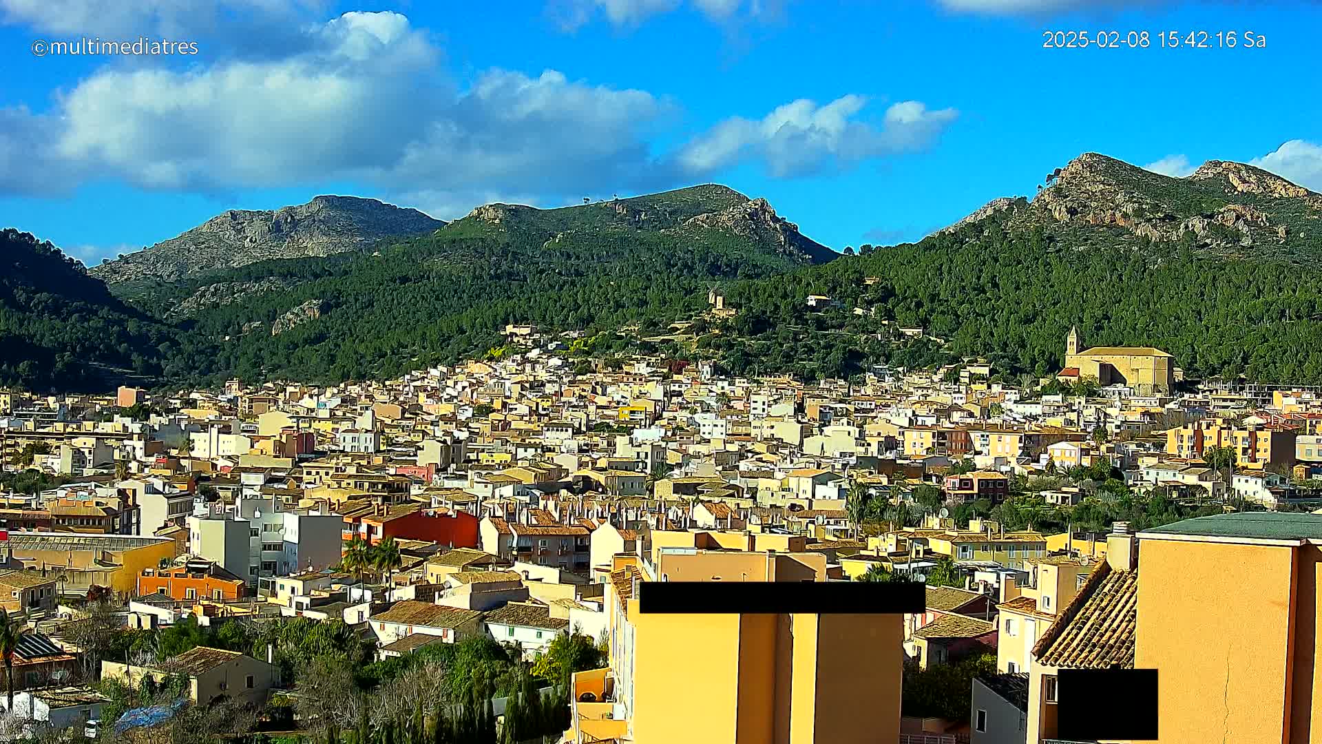 A sprawling town of low-rise buildings nestles in a valley surrounded by verdant, mountainous terrain under a mostly sunny sky with scattered clouds.