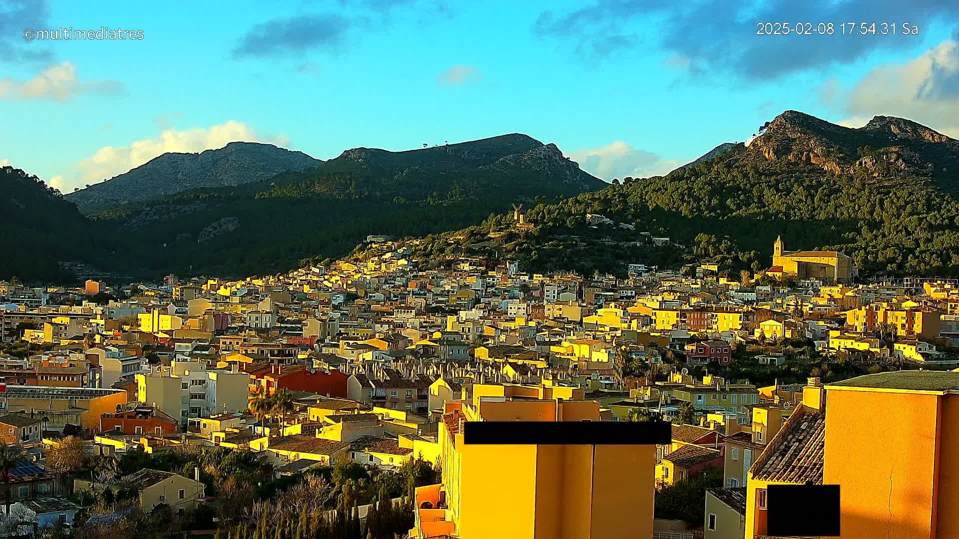 A dense collection of yellow-toned buildings sits nestled at the base of a mountainous landscape under a partly cloudy sunset sky.