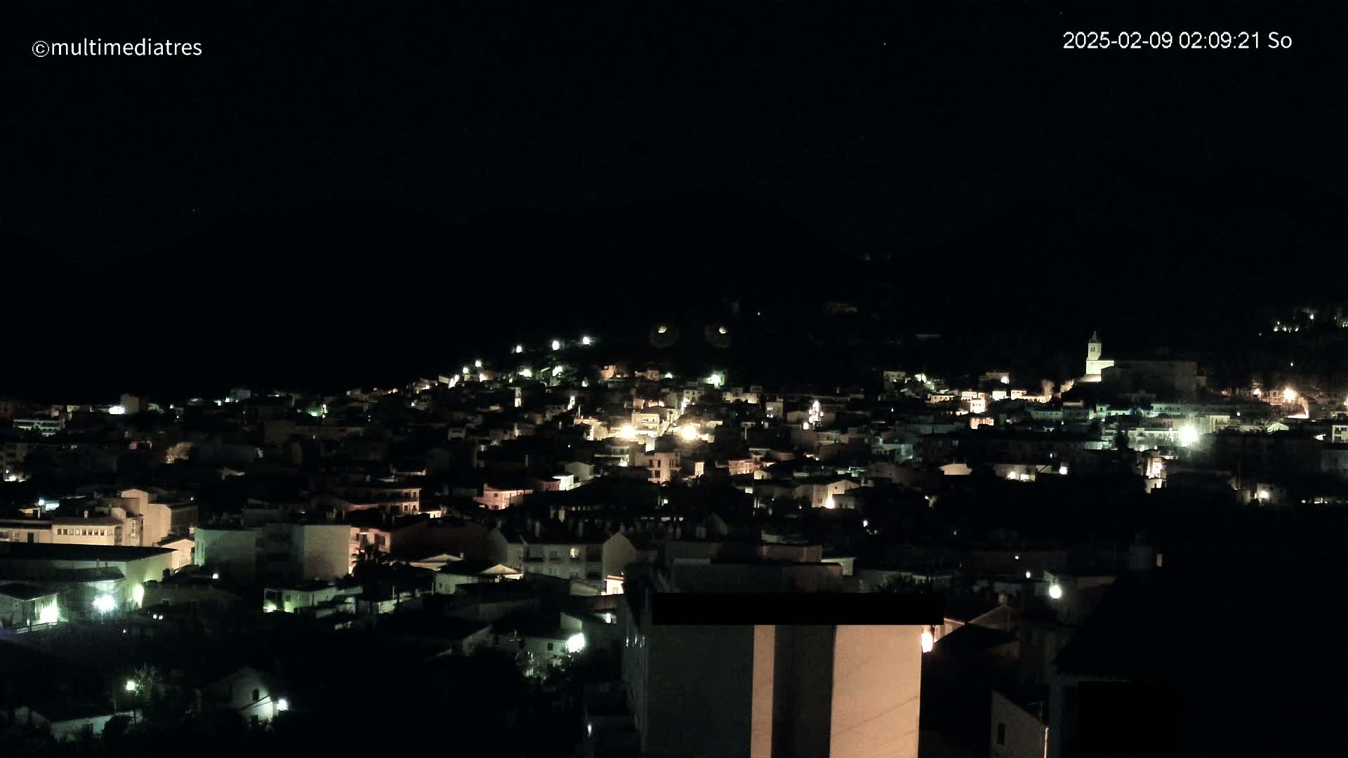A nighttime view of a town nestled on a hillside, illuminated by scattered lights under a clear, dark sky.