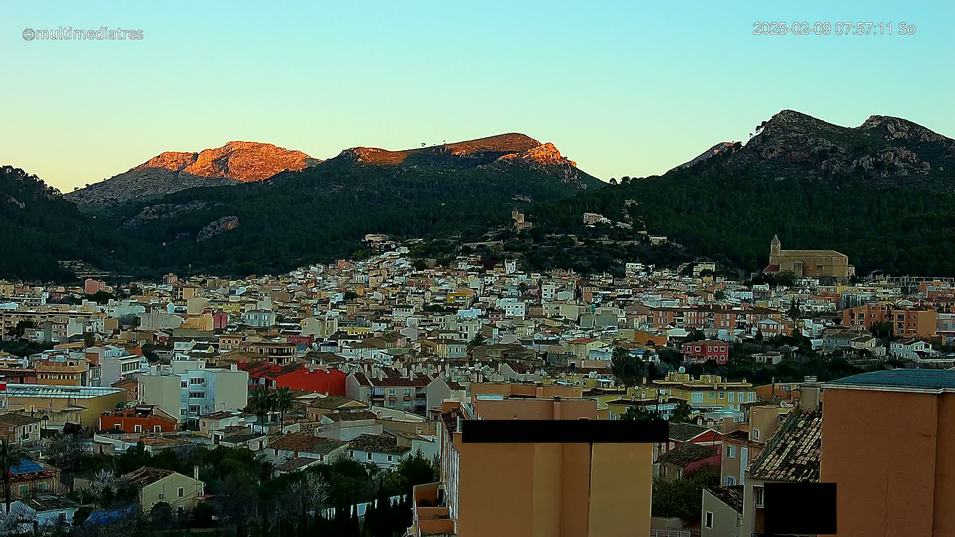 A sprawling town of low-rise buildings nestled at the base of sunlit, tree-covered mountains under a clear twilight sky.