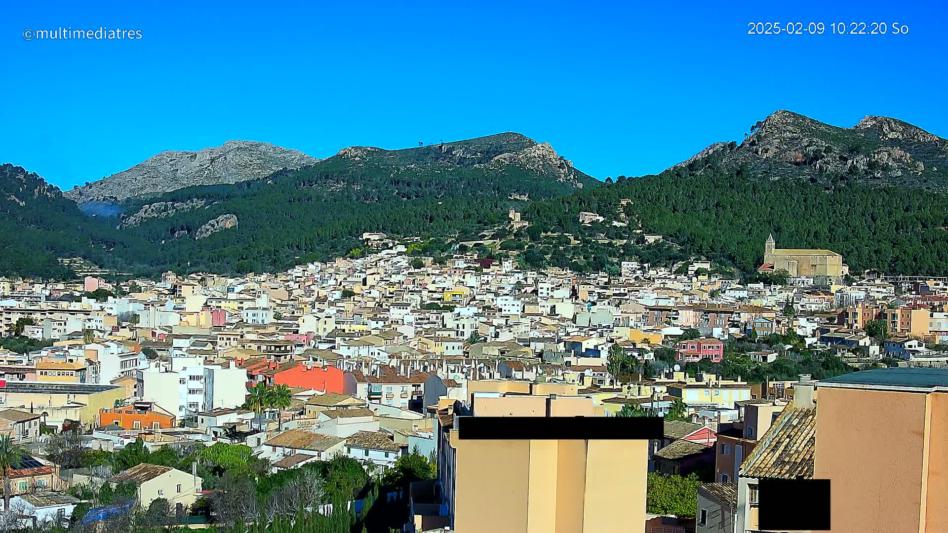A sprawling town of low-rise buildings nestled in a valley surrounded by green, tree-covered mountains under a clear blue sky.