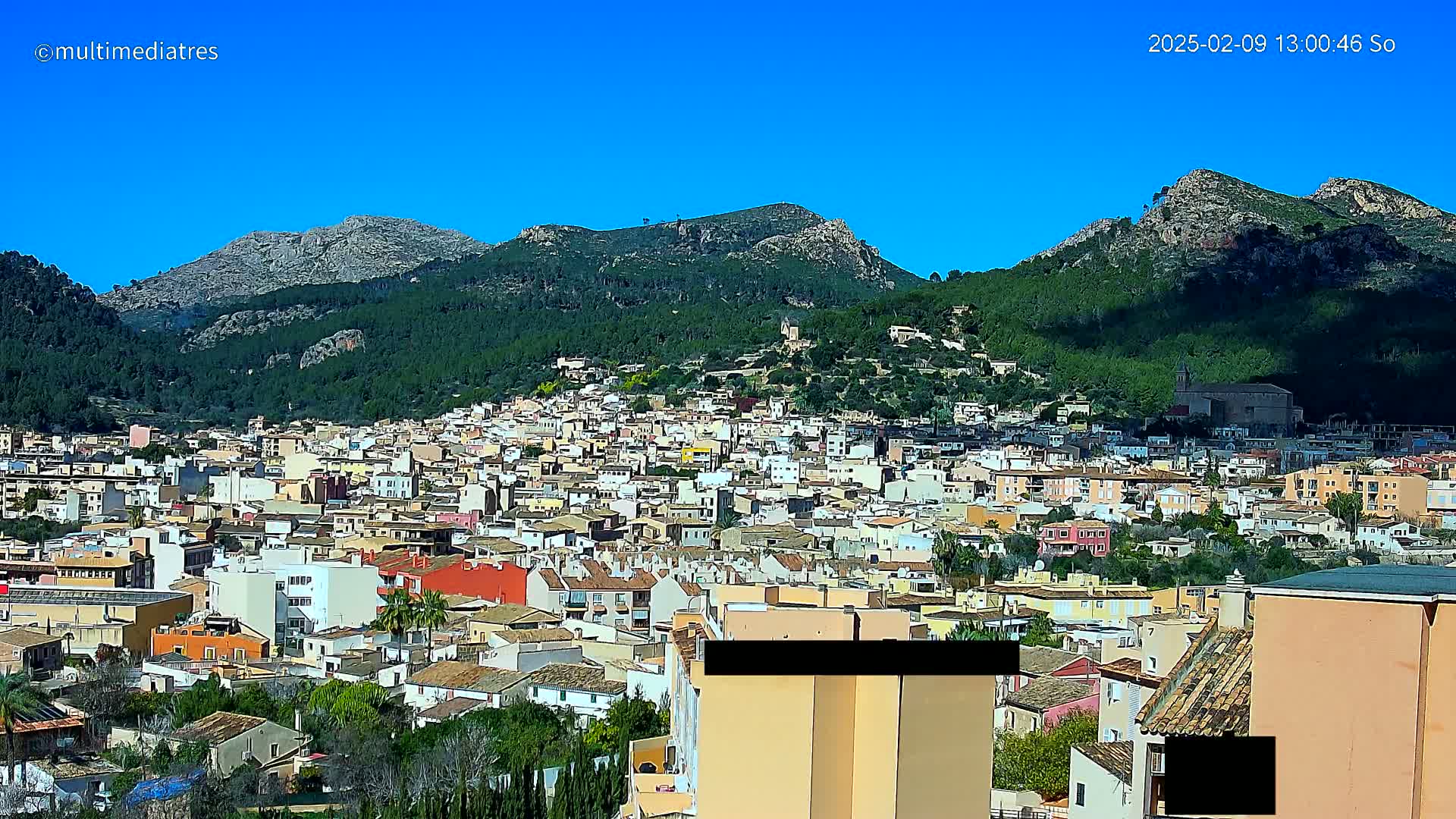 A sprawling town of low-rise buildings nestles against a backdrop of lush green mountains under a clear blue sky.