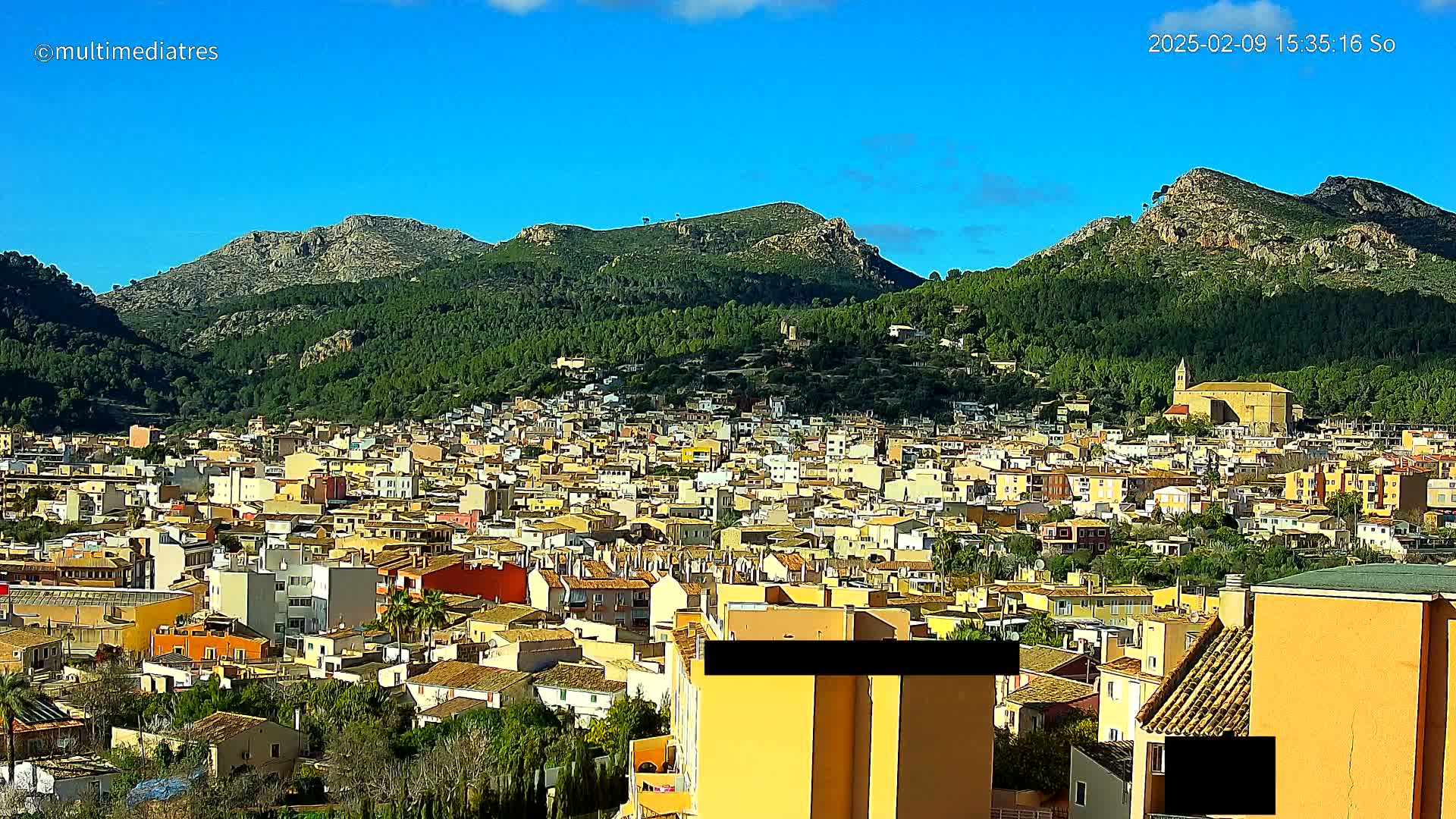 A sprawling town of low-rise buildings nestles at the foot of verdant, rocky mountains under a clear blue sky.