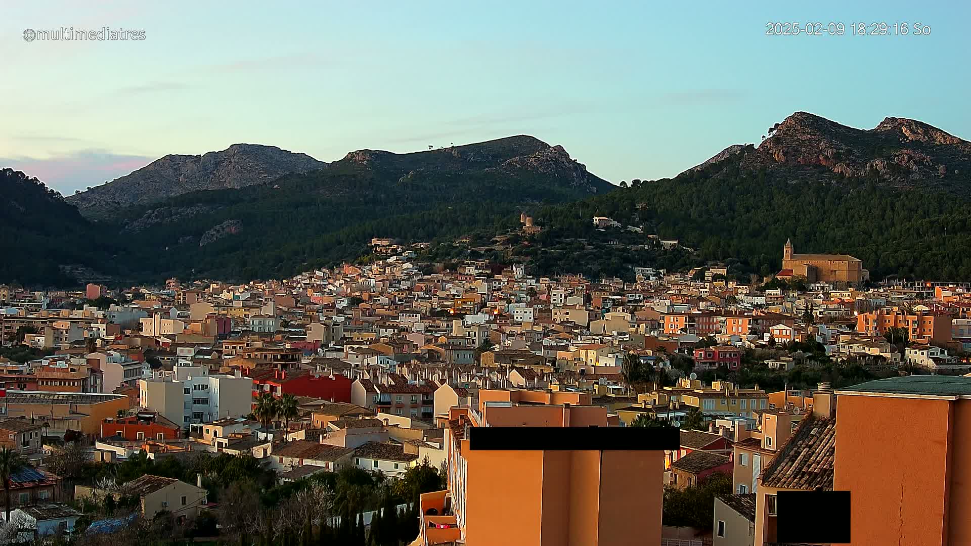 A sprawling town of low-rise buildings nestles at the foot of a mountain range under a clear twilight sky.