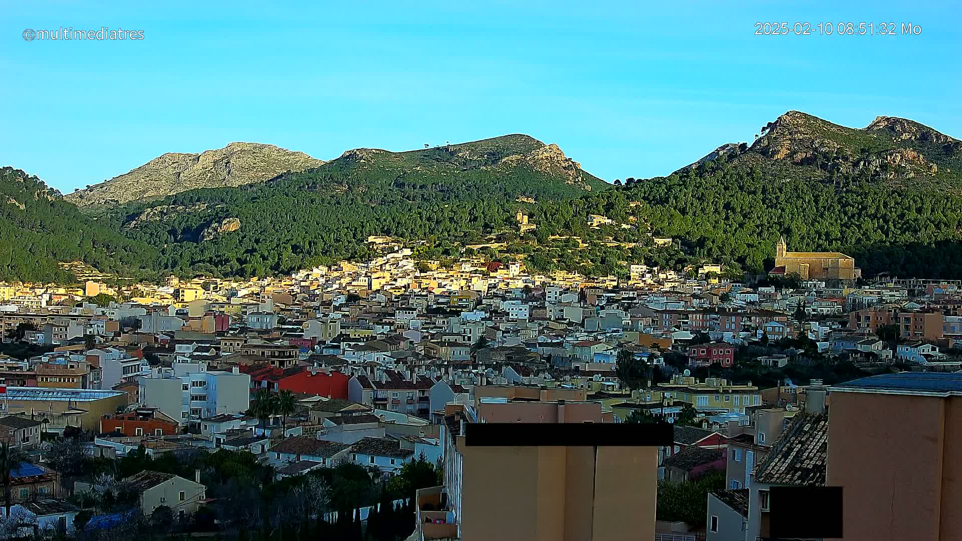 A sprawling town nestled in a valley between verdant, mountainous hills under a clear, bright blue sky.