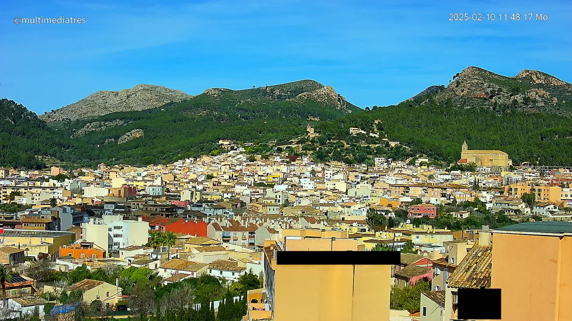 A sprawling town of low-rise buildings nestled in a valley between verdant, mountainous hills under a clear blue sky.
