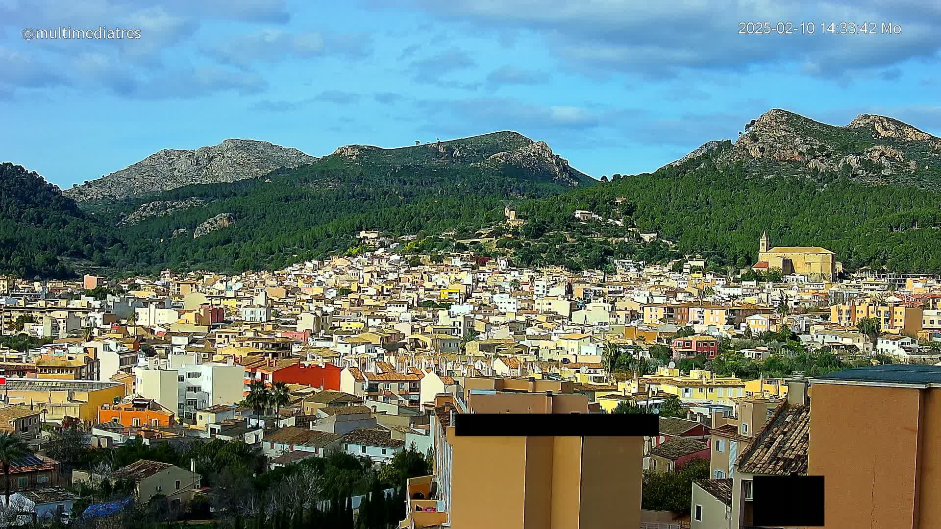A sprawling town of predominantly low-rise, light-colored buildings nestled in a valley surrounded by green, hilly terrain under a partly cloudy sky.