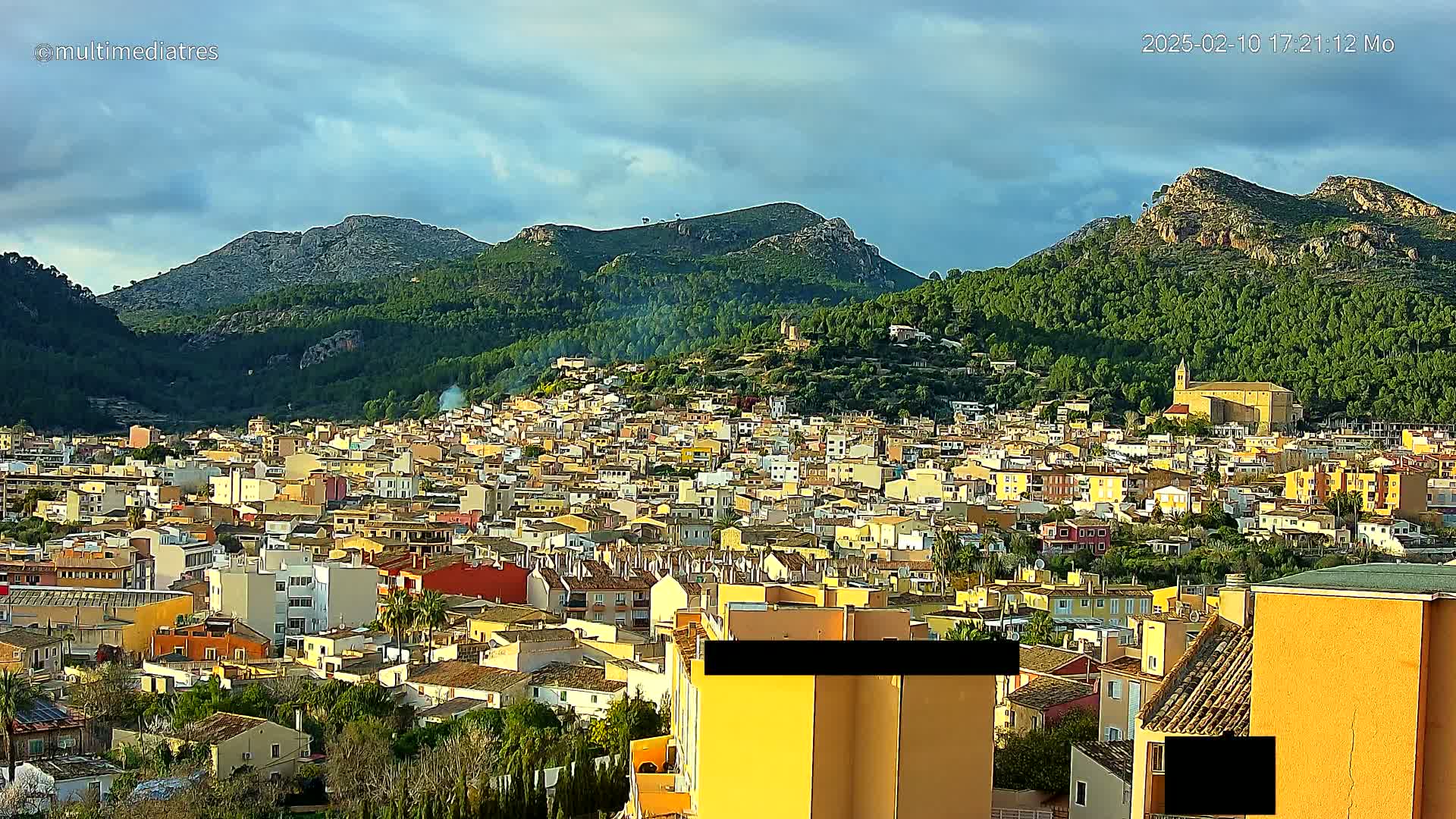 A dense collection of low-rise buildings stretches across a hillside, nestled beneath mountains under a partly cloudy sky.